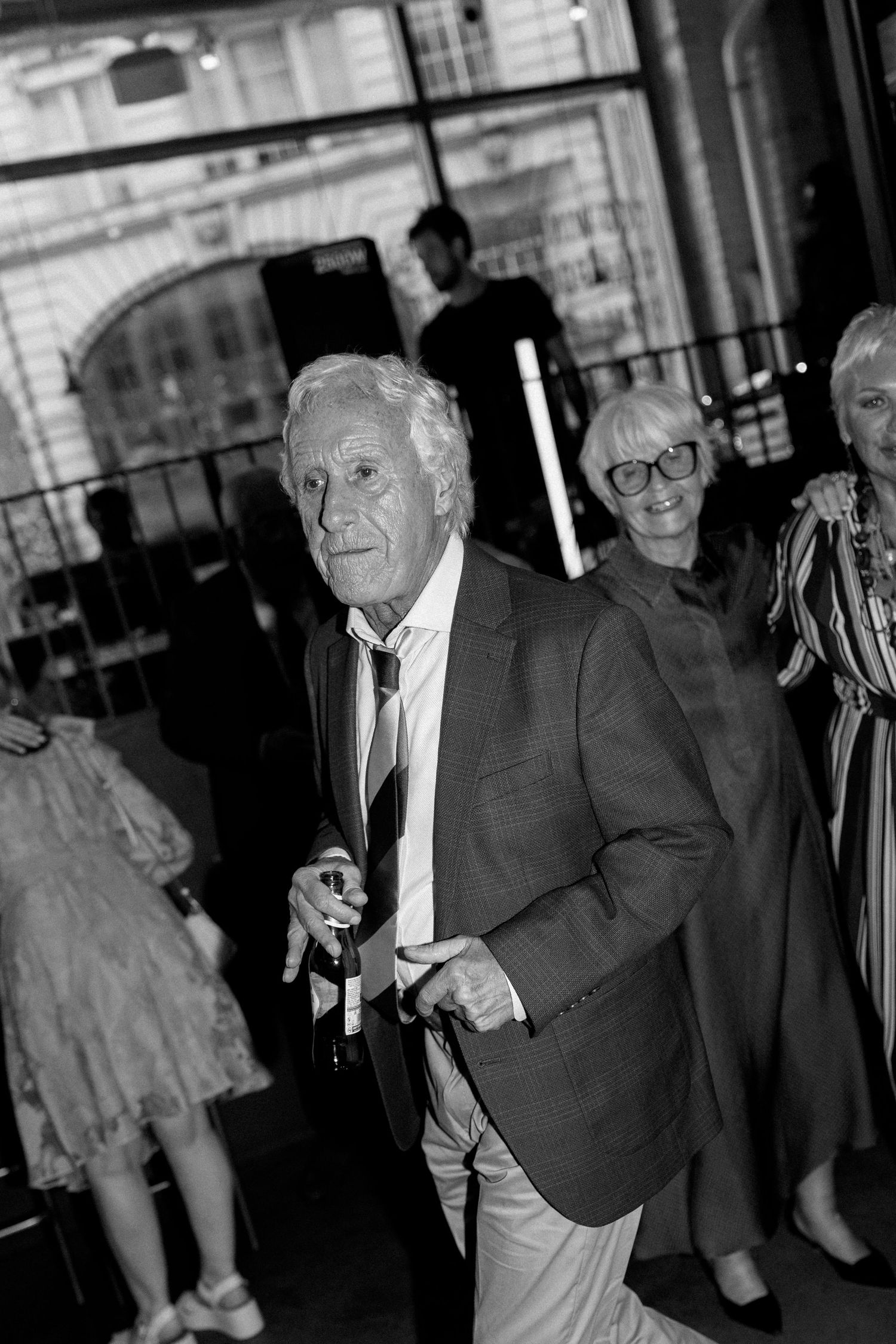 Elderly gentleman in suit with striped tie holds bottle at elegant indoor wedding reception venue.