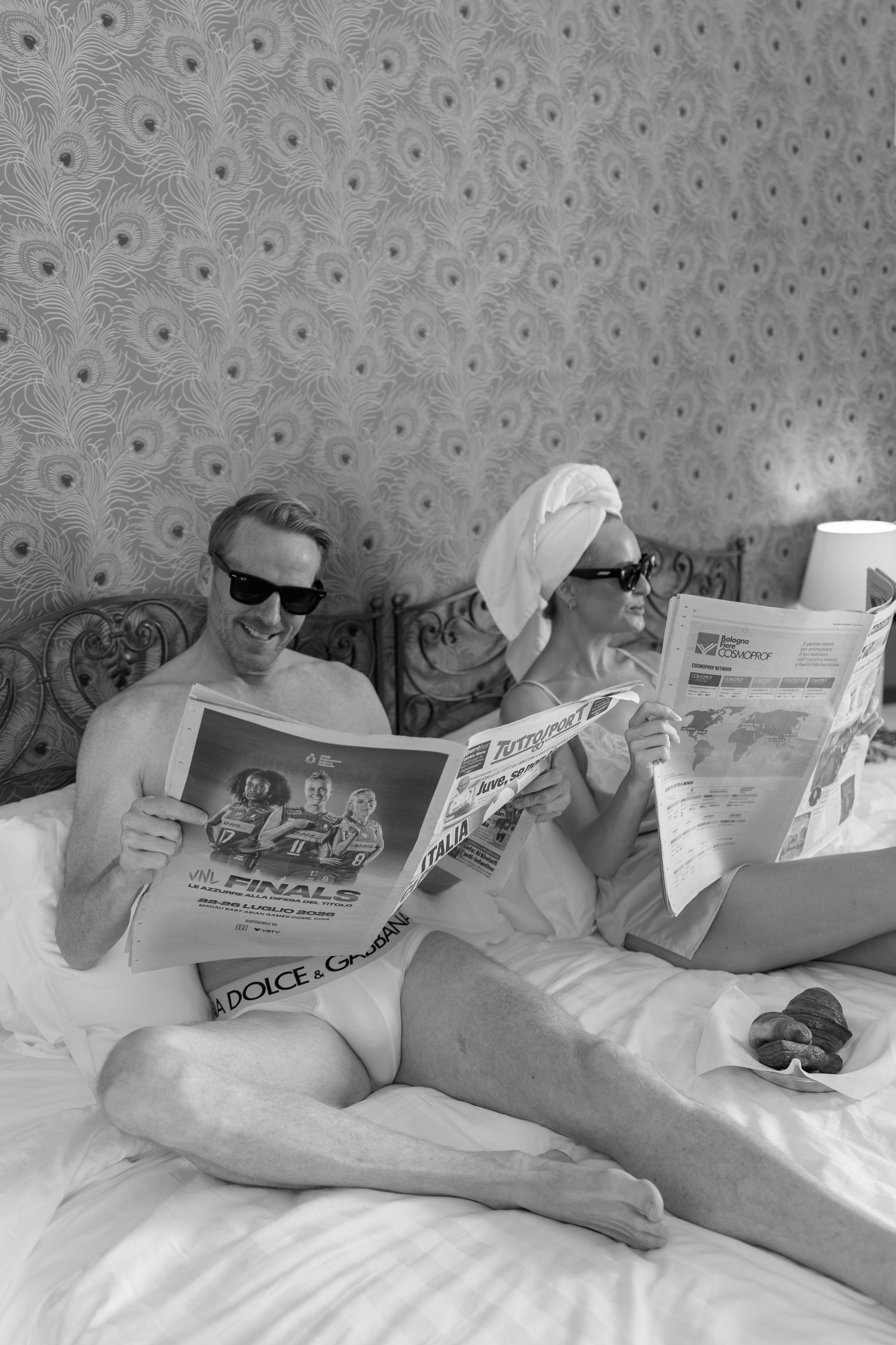 Black and white photo of couple relaxing in bed reading magazines and newspapers, wearing sunglasses and towel turban.