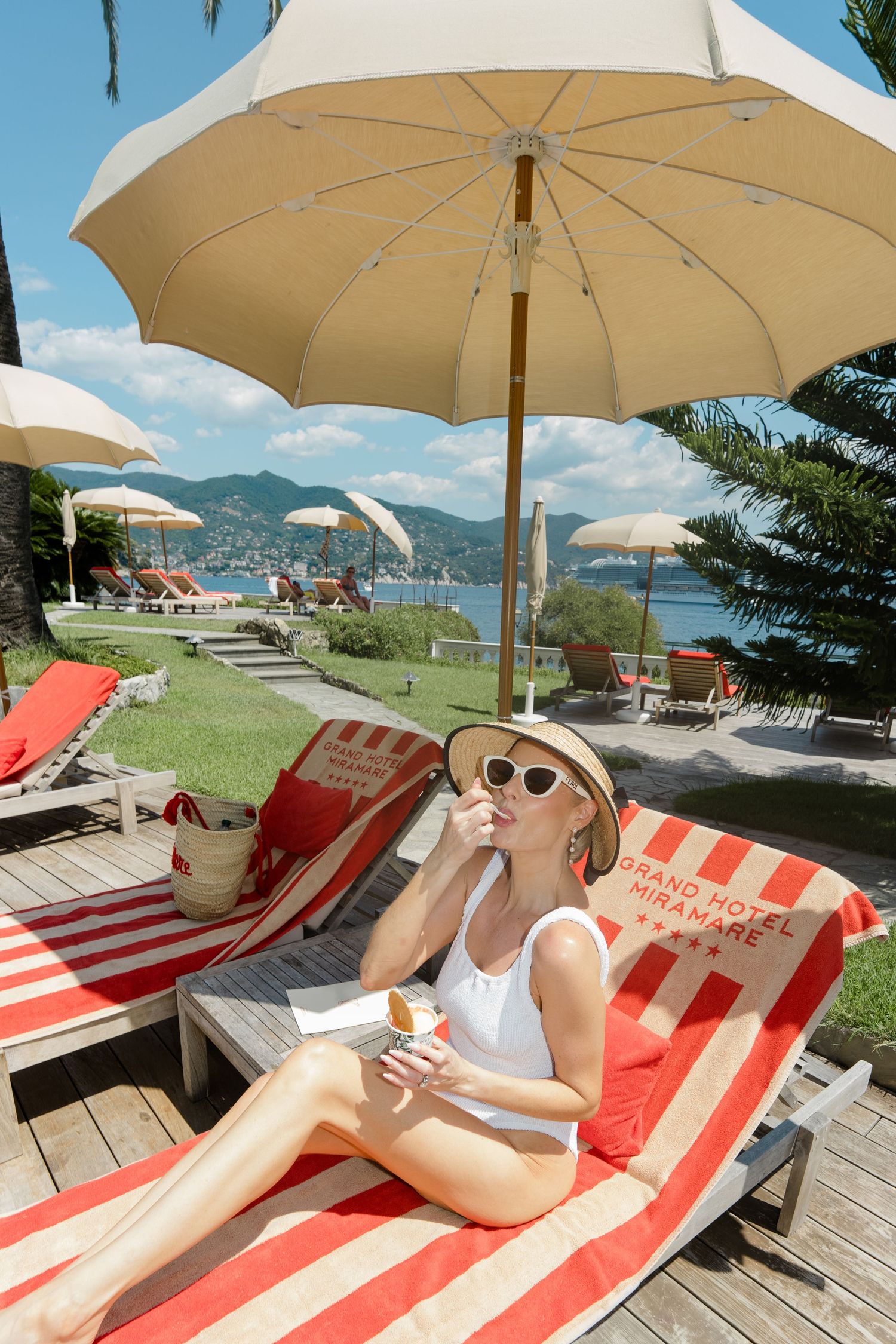 Elegant woman in white swimsuit relaxes on striped Grand Hotel Miramare lounger under umbrella eating gelato.