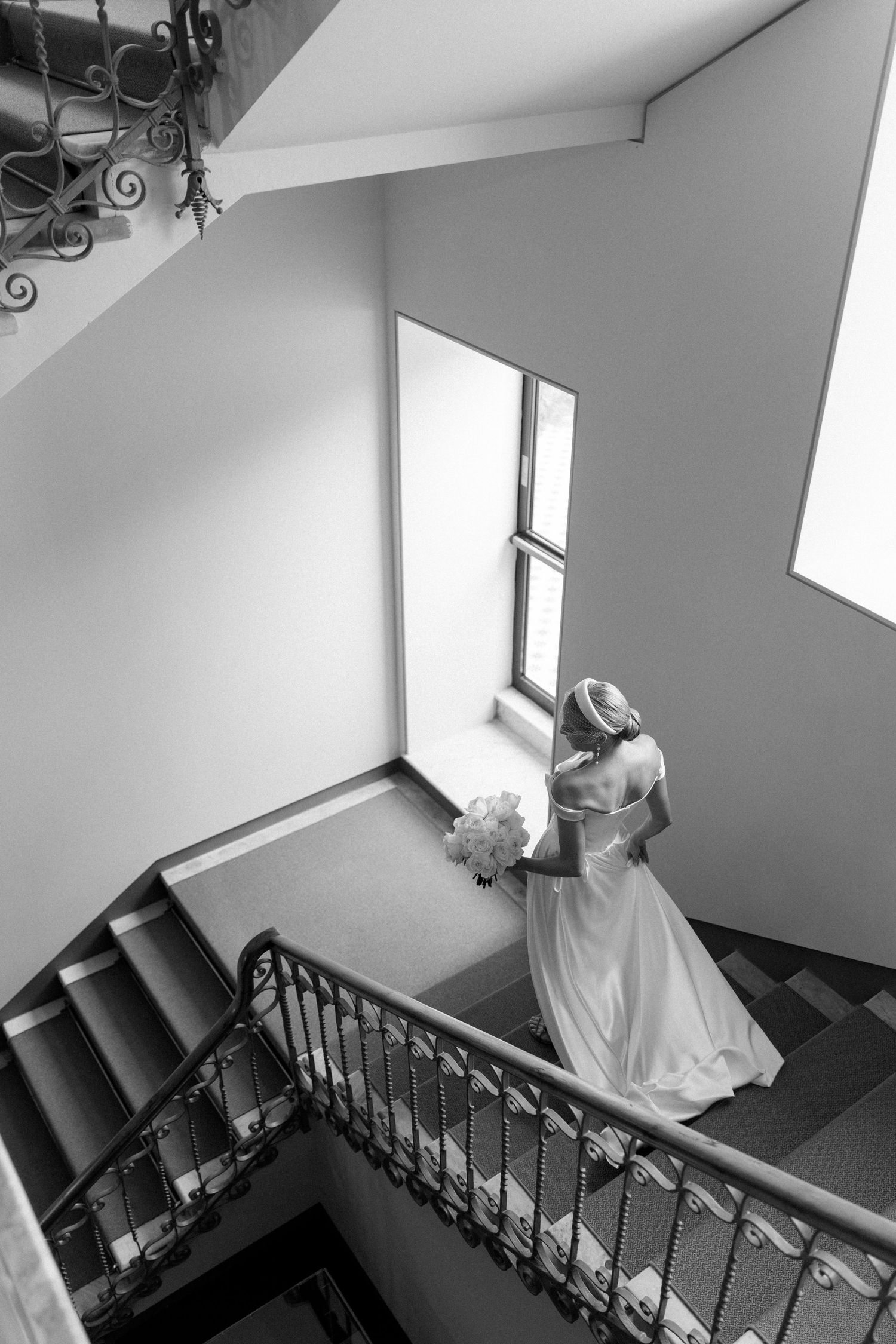 Bride in white gown holding bouquet on grand staircase, black and white wedding photo.
