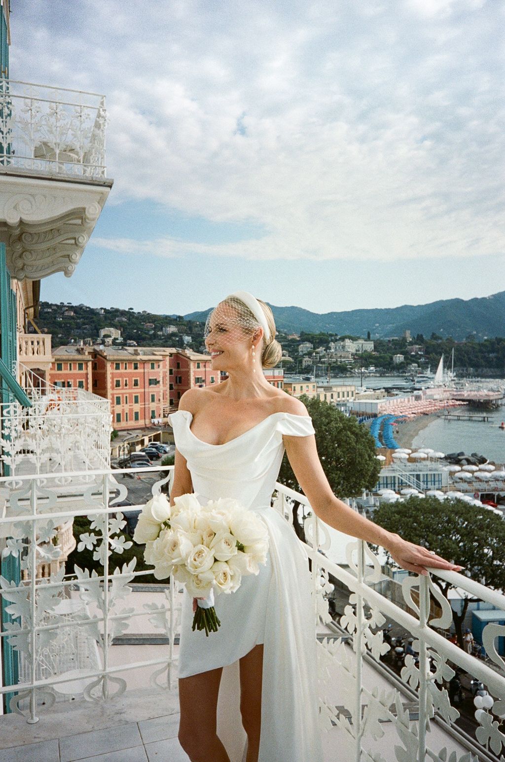 Smiling bride in white off-shoulder wedding dress with bouquet on balcony overlooking harbor.