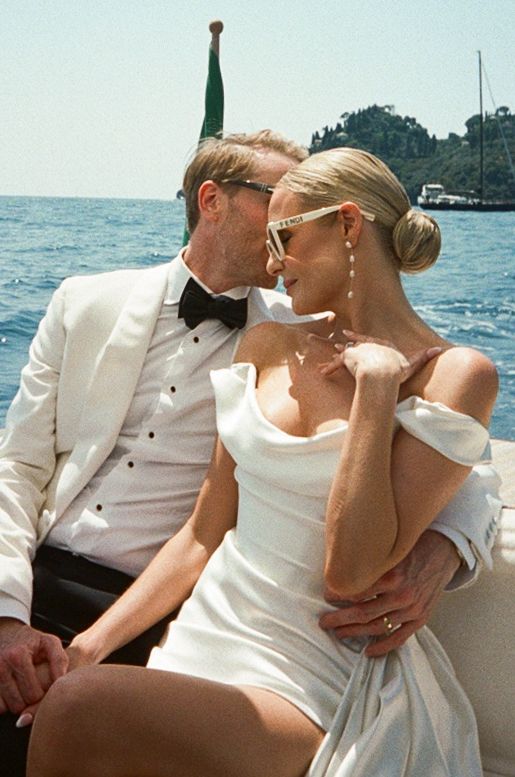 Elegant couple in wedding attire on a boat, groom in white tuxedo and bride in off-shoulder dress, Mediterranean backdrop.