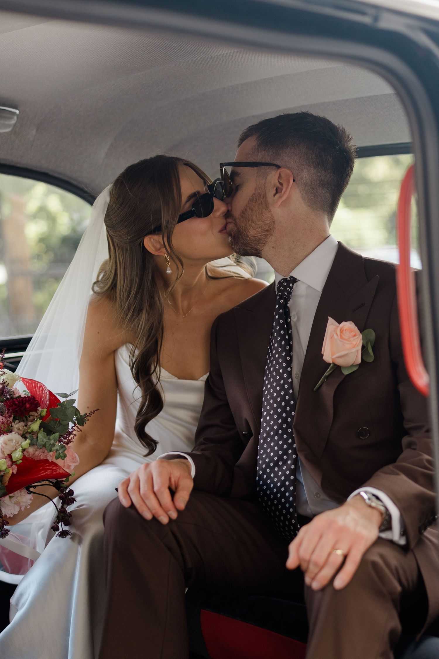Bride and groom kissing in vintage car, wearing sunglasses, bride holding red floral bouquet.