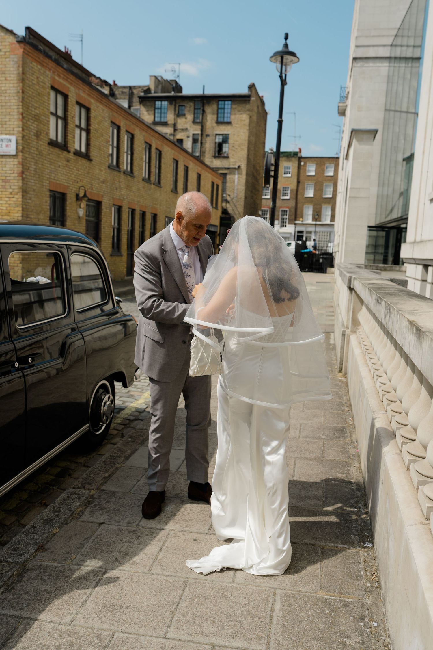 Man helping bride adjust her white wedding veil on a sunny London street.