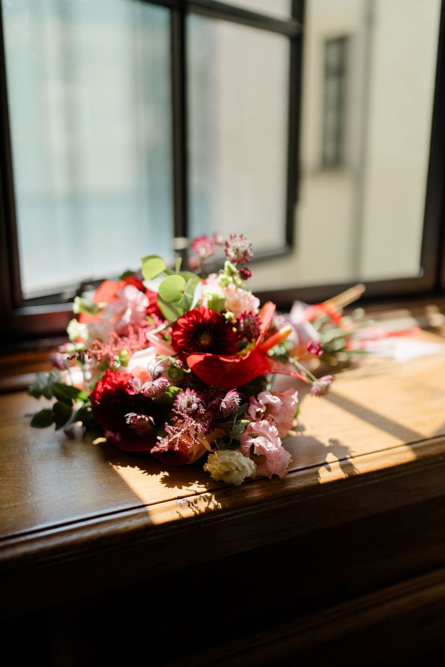 Vibrant red and pink wedding bouquet resting on wooden windowsill bathed in natural sunlight.