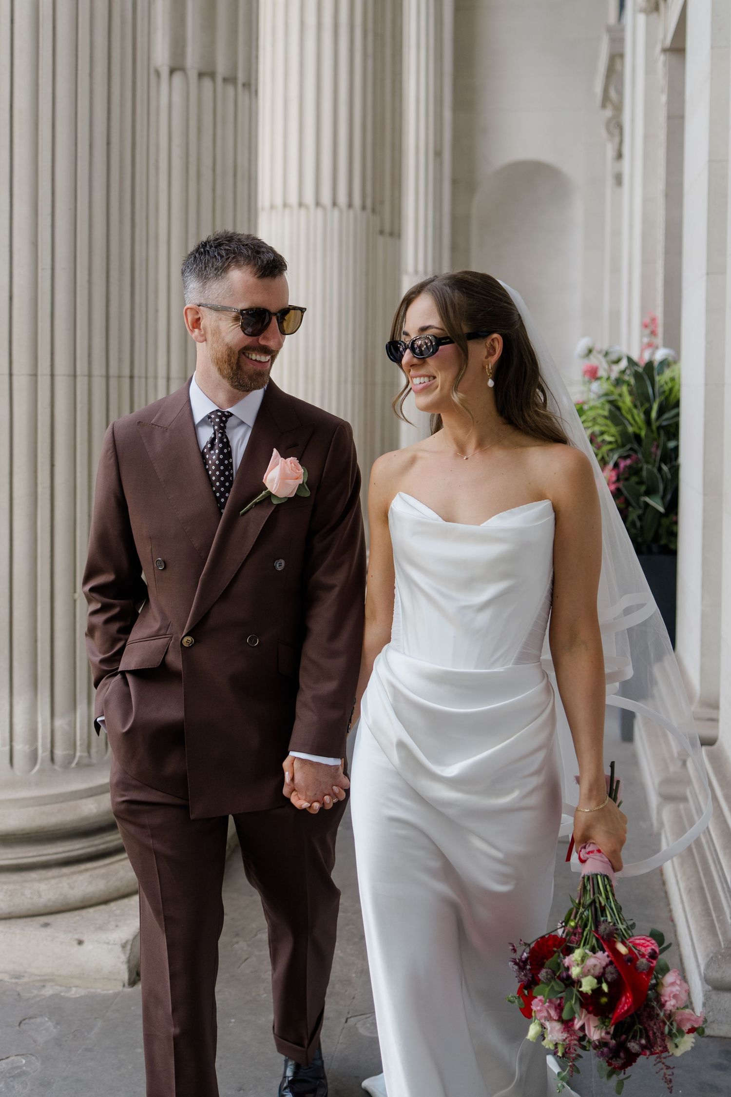 Stylish newlyweds walking hand in hand wearing sunglasses, groom in brown double-breasted suit, bride in strapless white gown.
