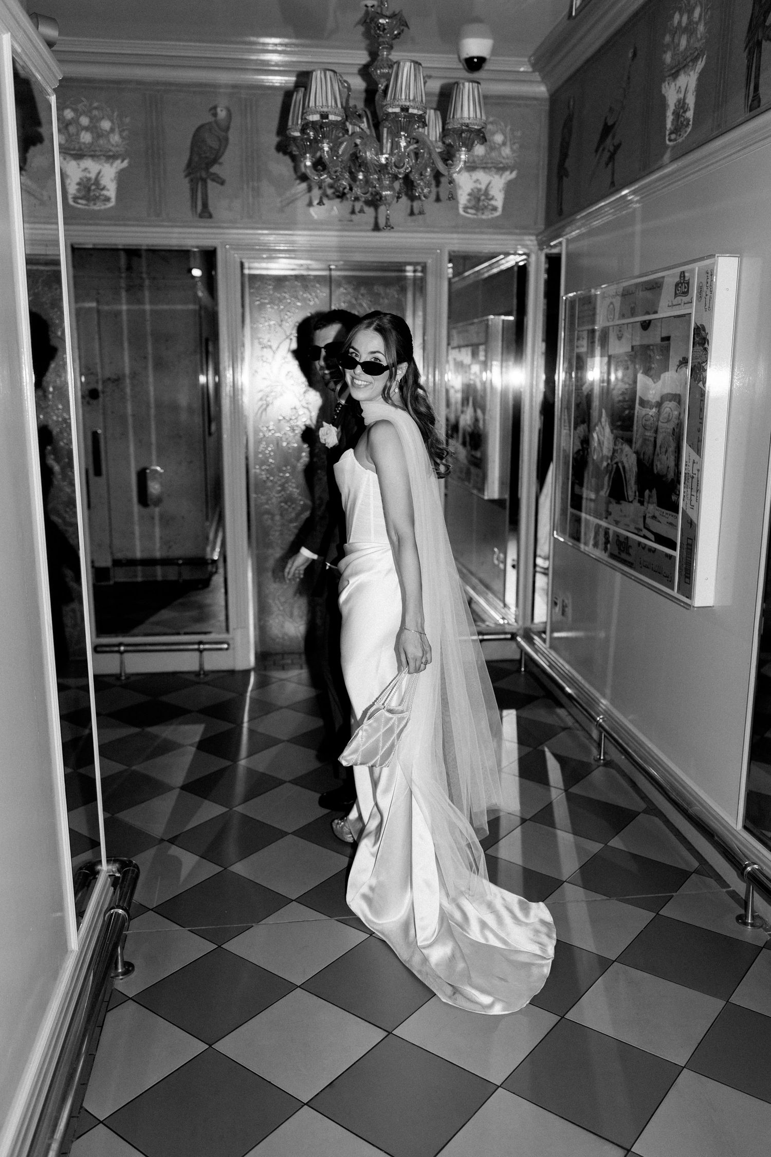 Black and white photo of bride in sleek wedding dress and veil posing in ornate hotel corridor.