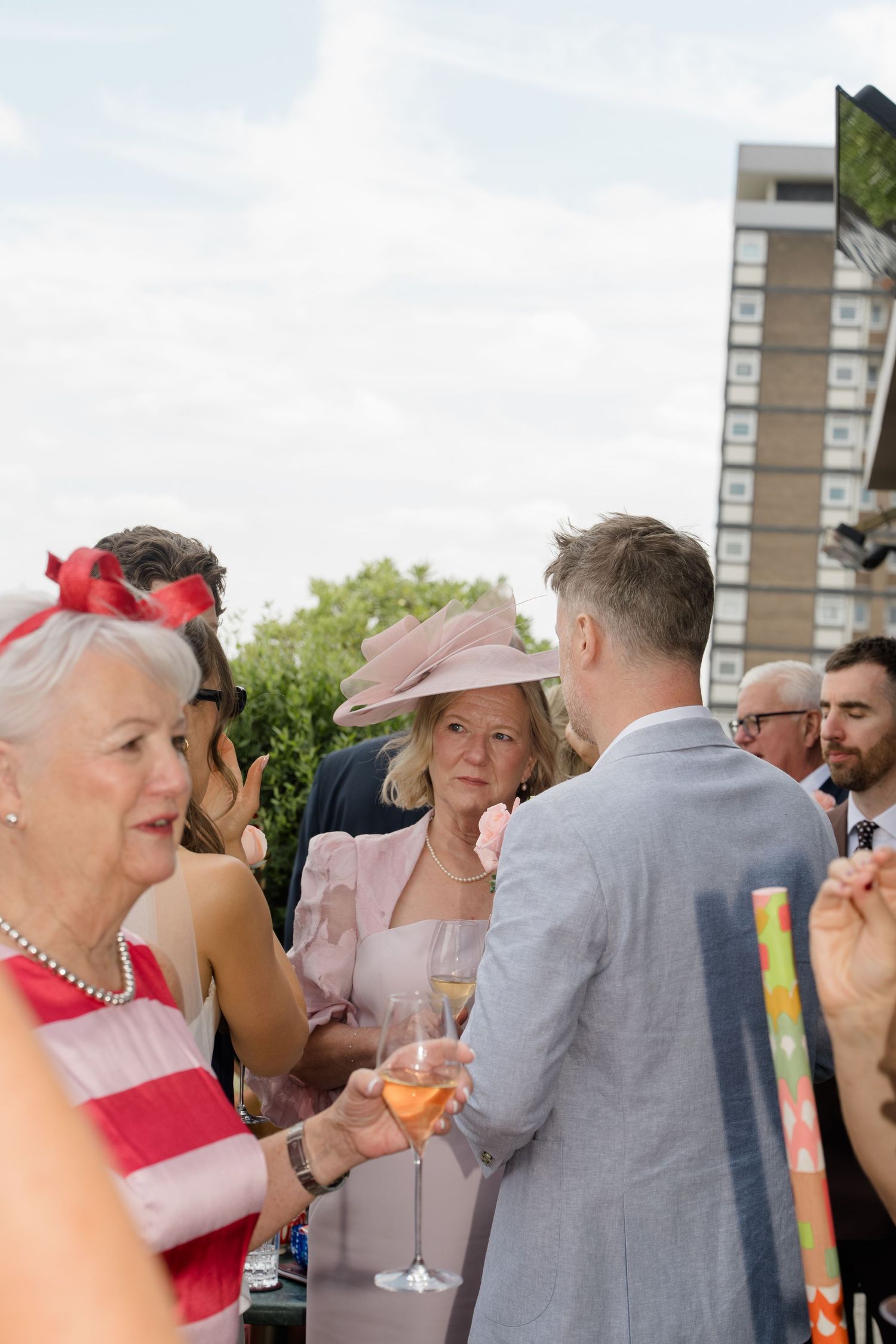 Wedding guests mingle outdoors, women wearing elegant hats and holding champagne glasses at reception.
