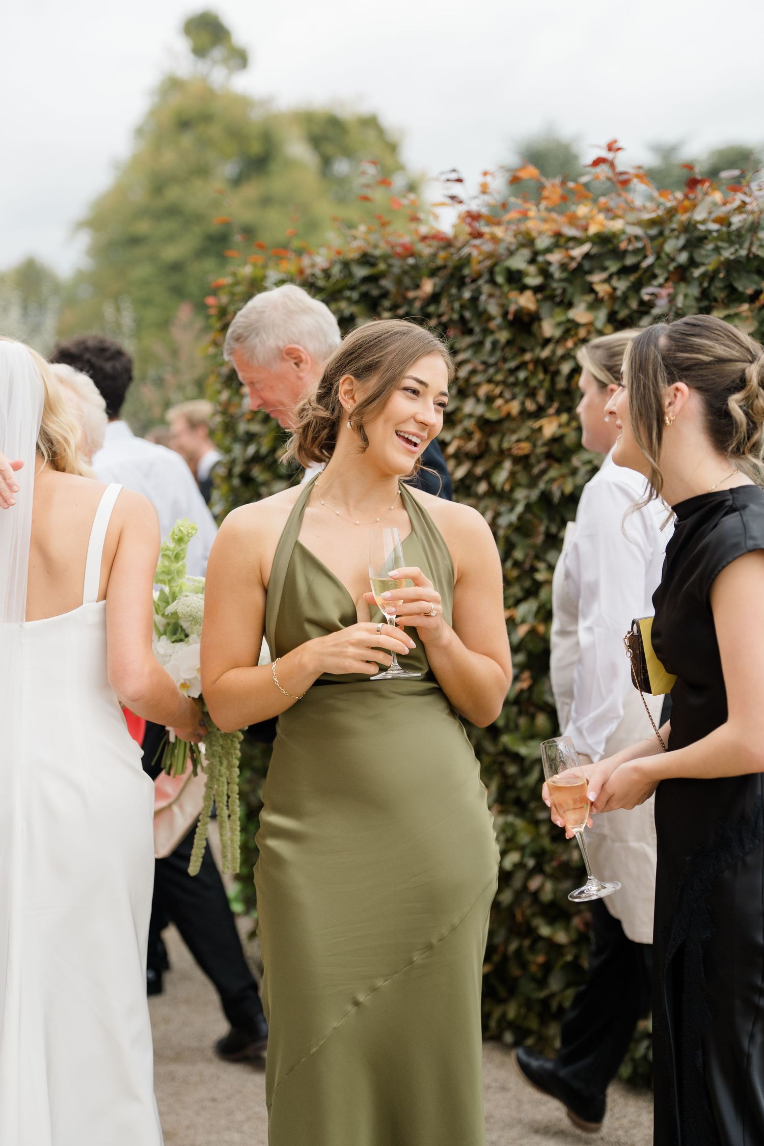 Bridesmaid in olive green halter dress laughs while holding champagne at outdoor wedding reception.