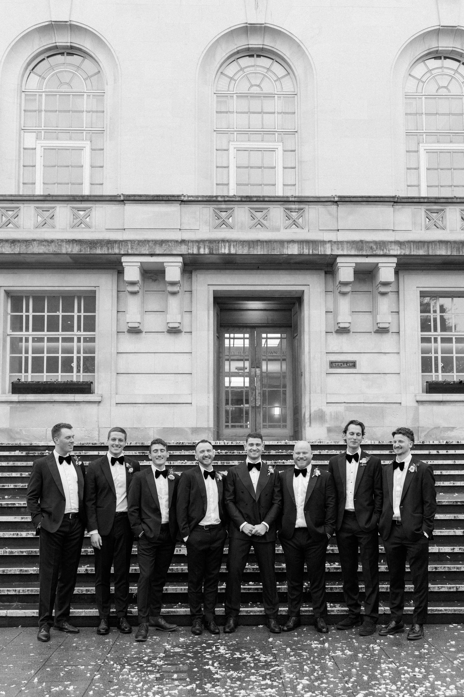 A row of formally dressed men in black tuxedos and bowties stand against a stone building exterior.