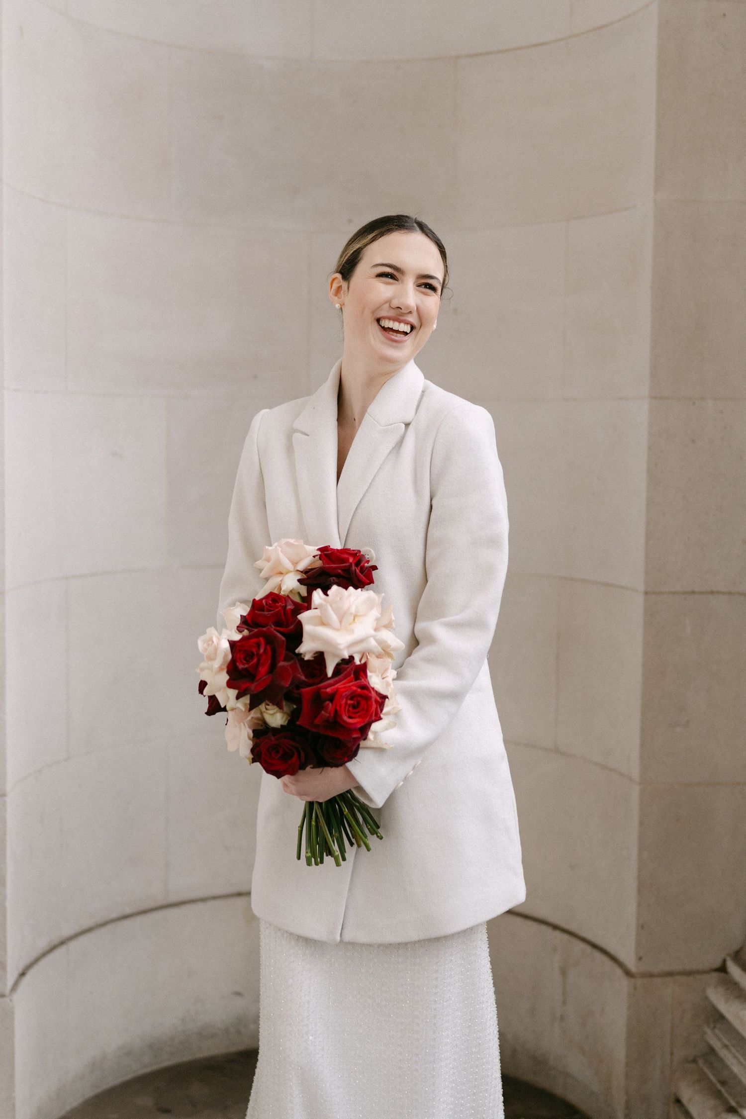A woman in a white blazer holds a stunning bouquet of red and white flowers against a stone wall.