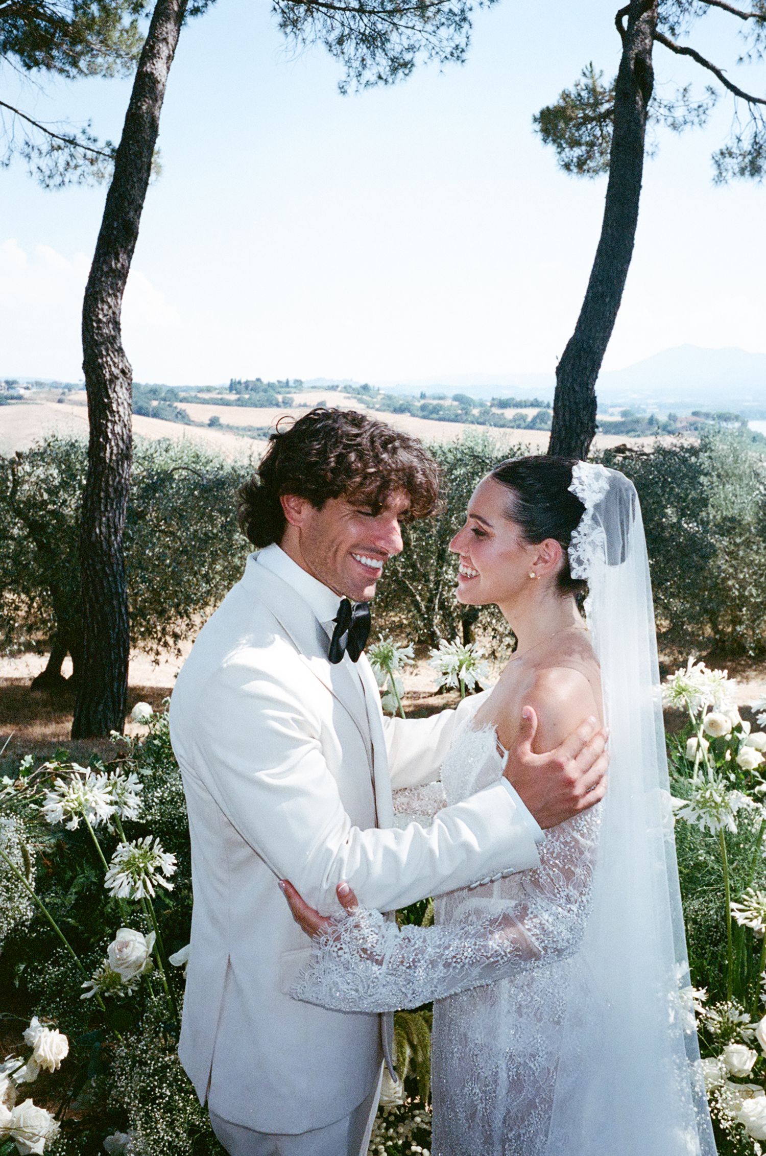 A romantic wedding couple shares a joyful moment together under pine trees in an outdoor garden setting.
