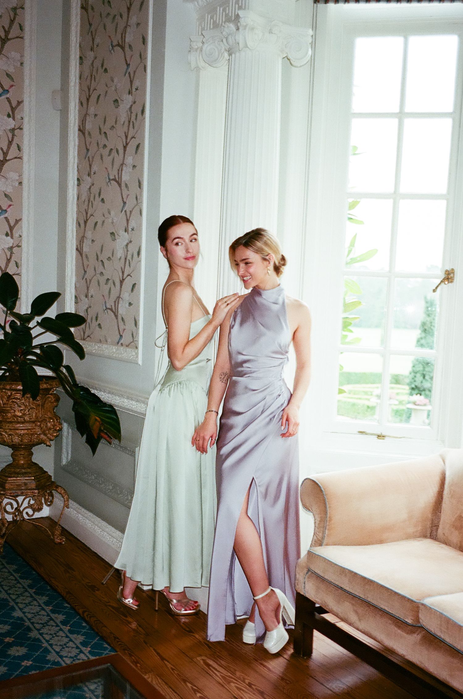 Two bridesmaids in lavender dresses help each other get ready in an elegant room with floral wallpaper.