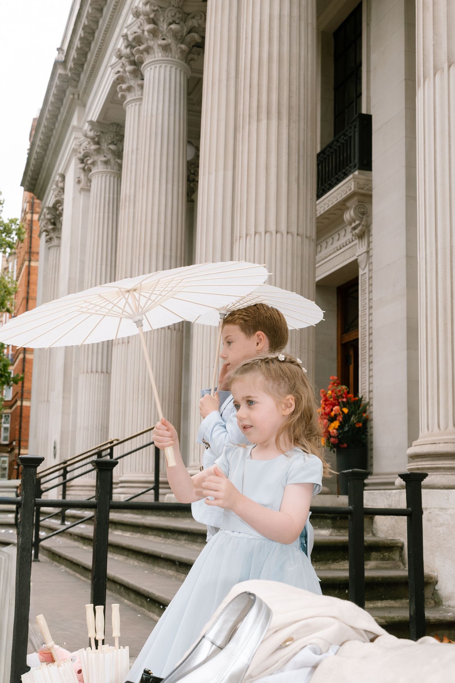 Young flower girls with white parasols pose along columned building exterior.