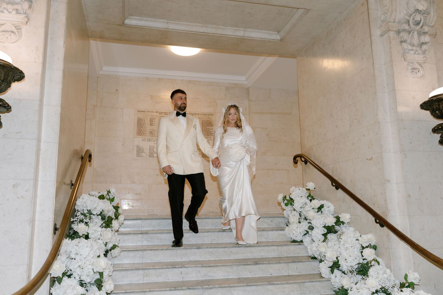 Wedding party walking down marble steps decorated with white floral arrangements.
