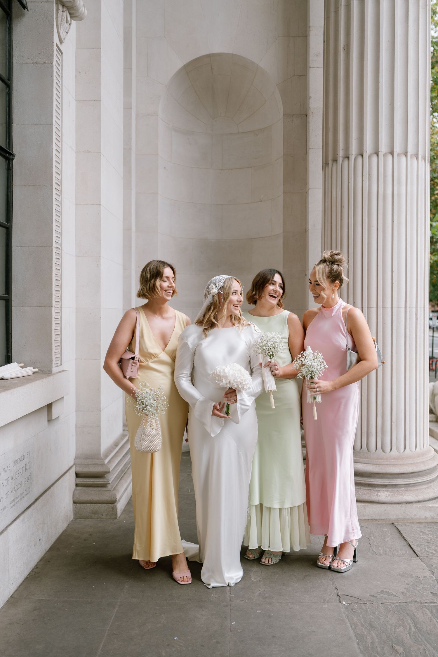 Bridesmaids in soft pastel dresses gathered against elegant white pillars.