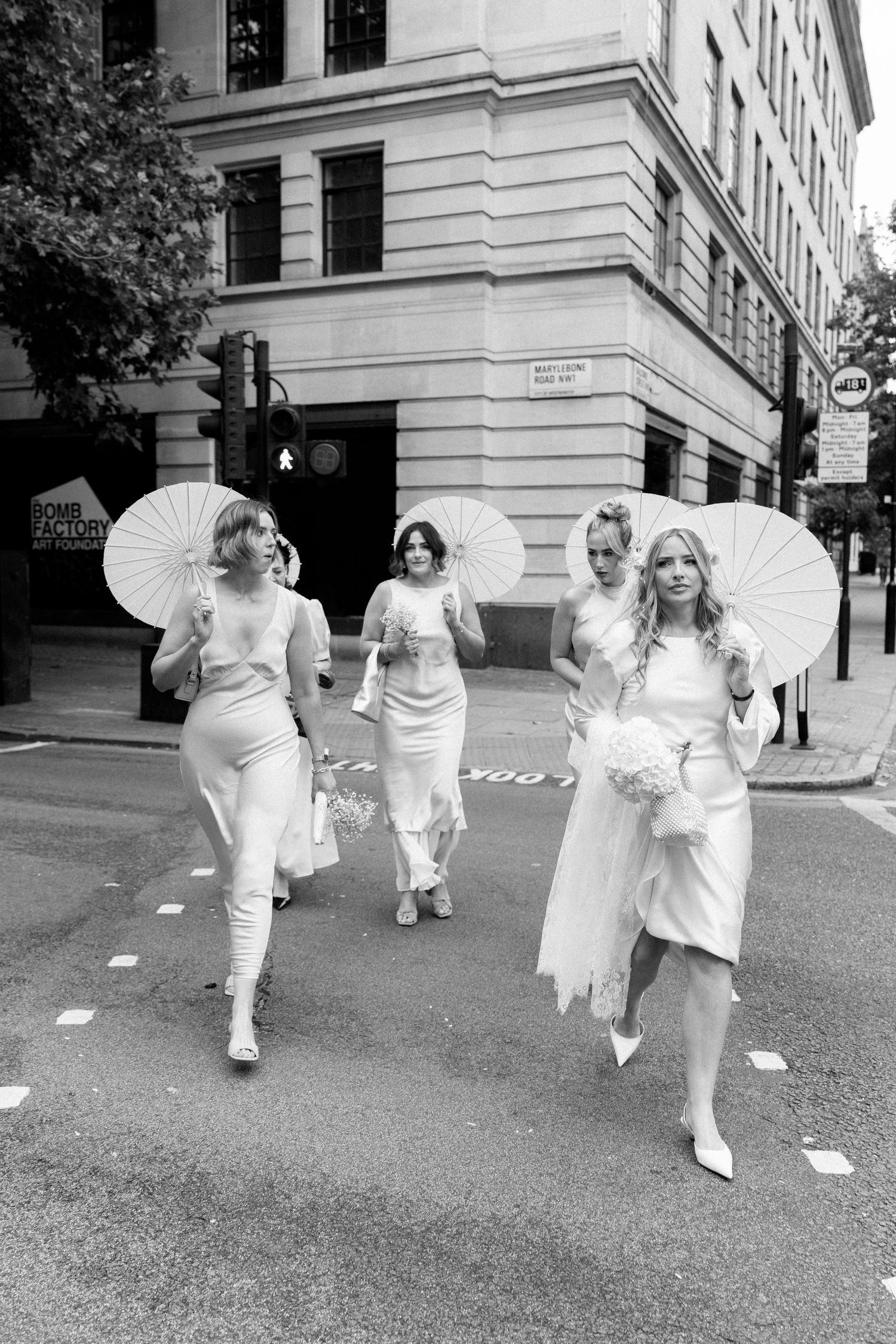 A wedding party walks down a city street carrying white parasols in elegant white dresses.