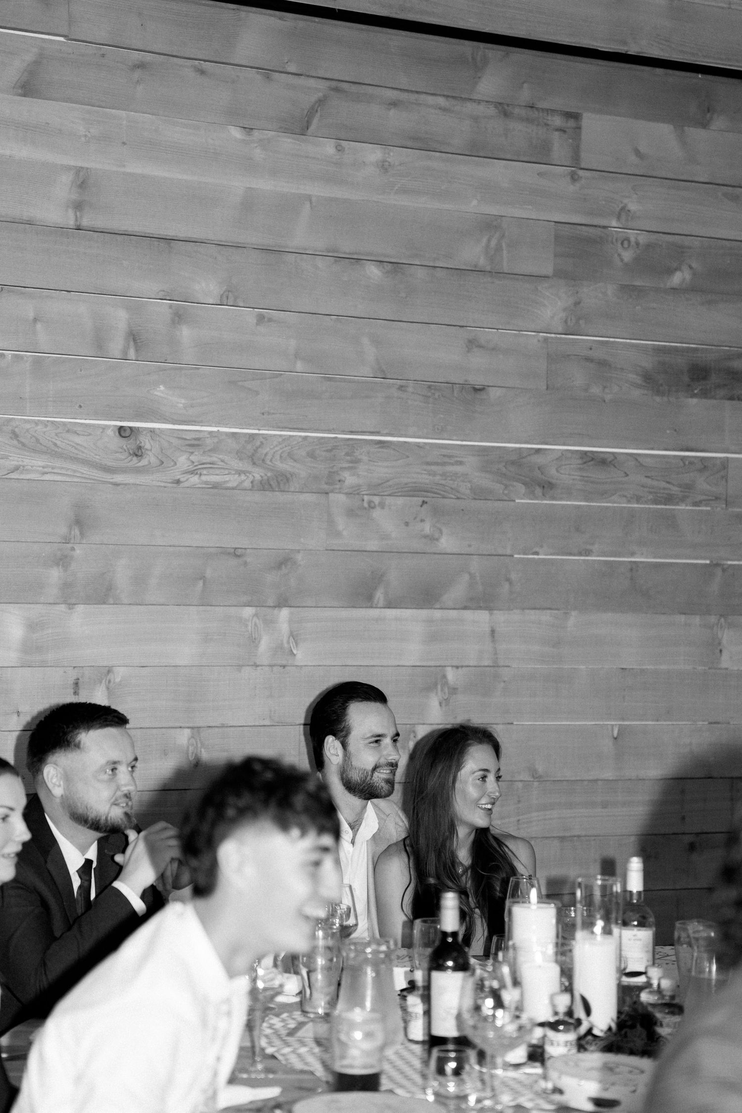 Wedding reception guests seated at long tables with drinks in rustic wooden venue.