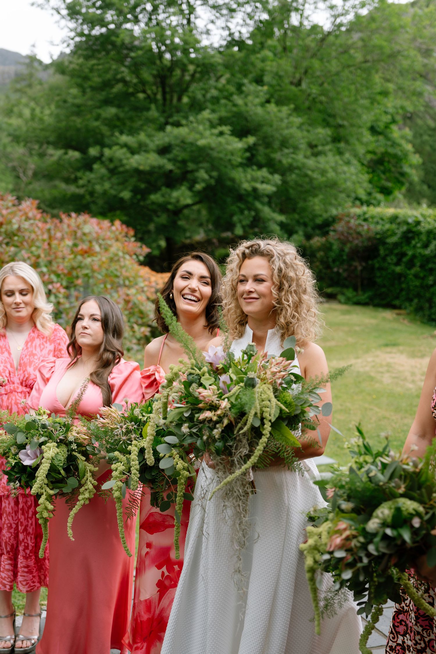 Bridal party in coral and white dresses holding greenery bouquets in a garden setting.