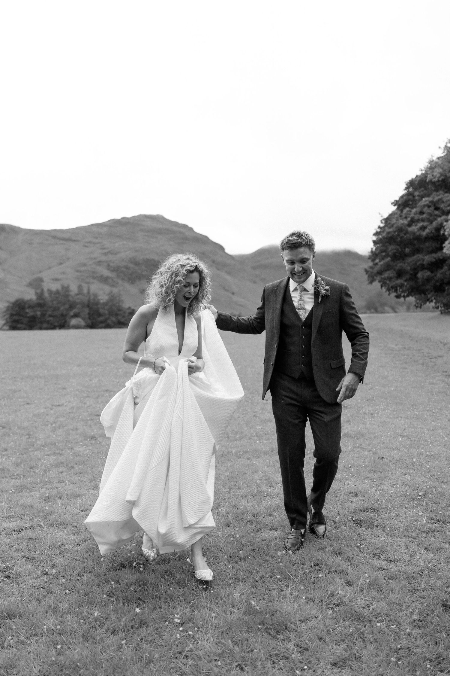 Wedding couple runs playfully across a green field with mountains in the distance.
