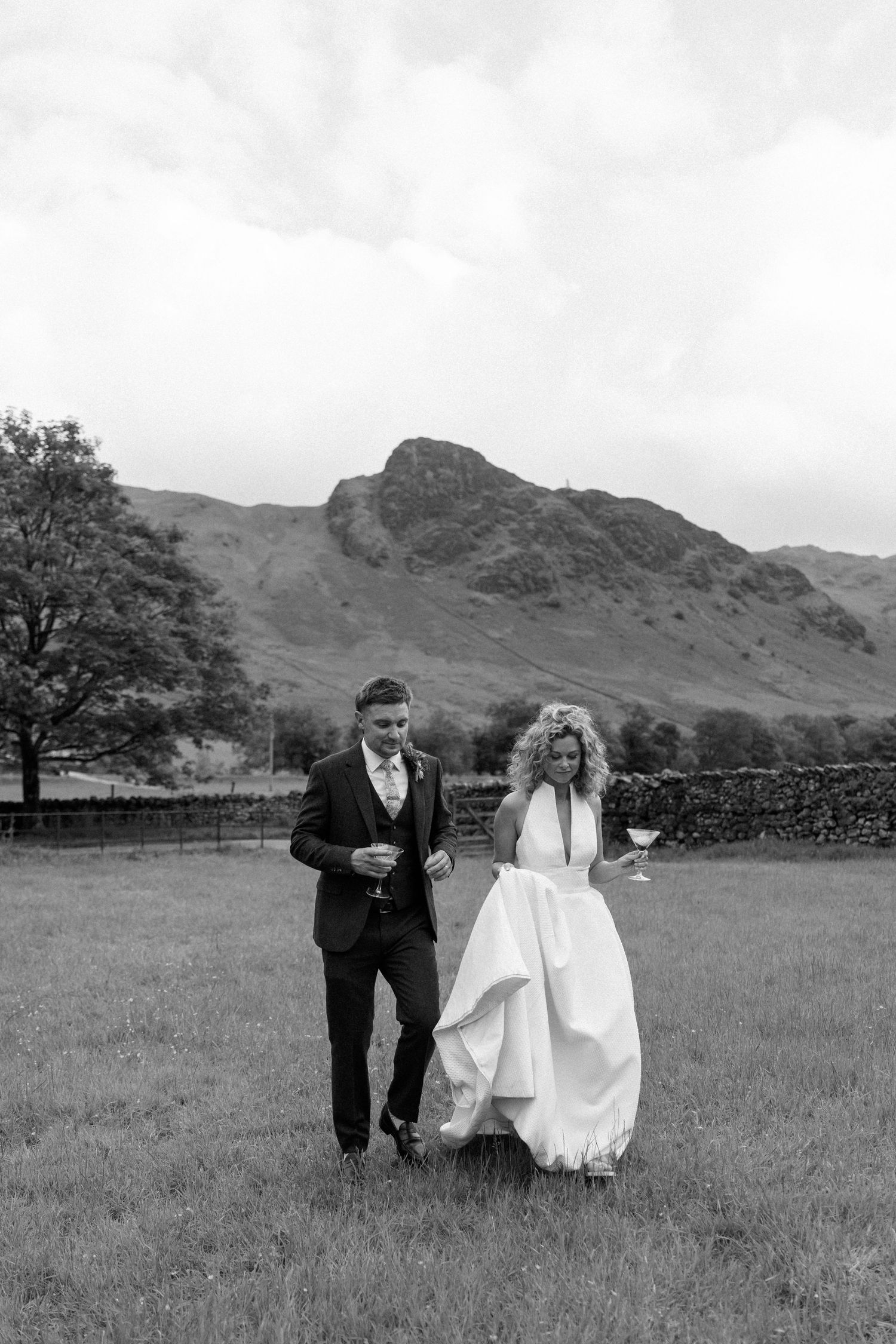 A couple runs playfully across a green field with mountains in the background during their wedding day.