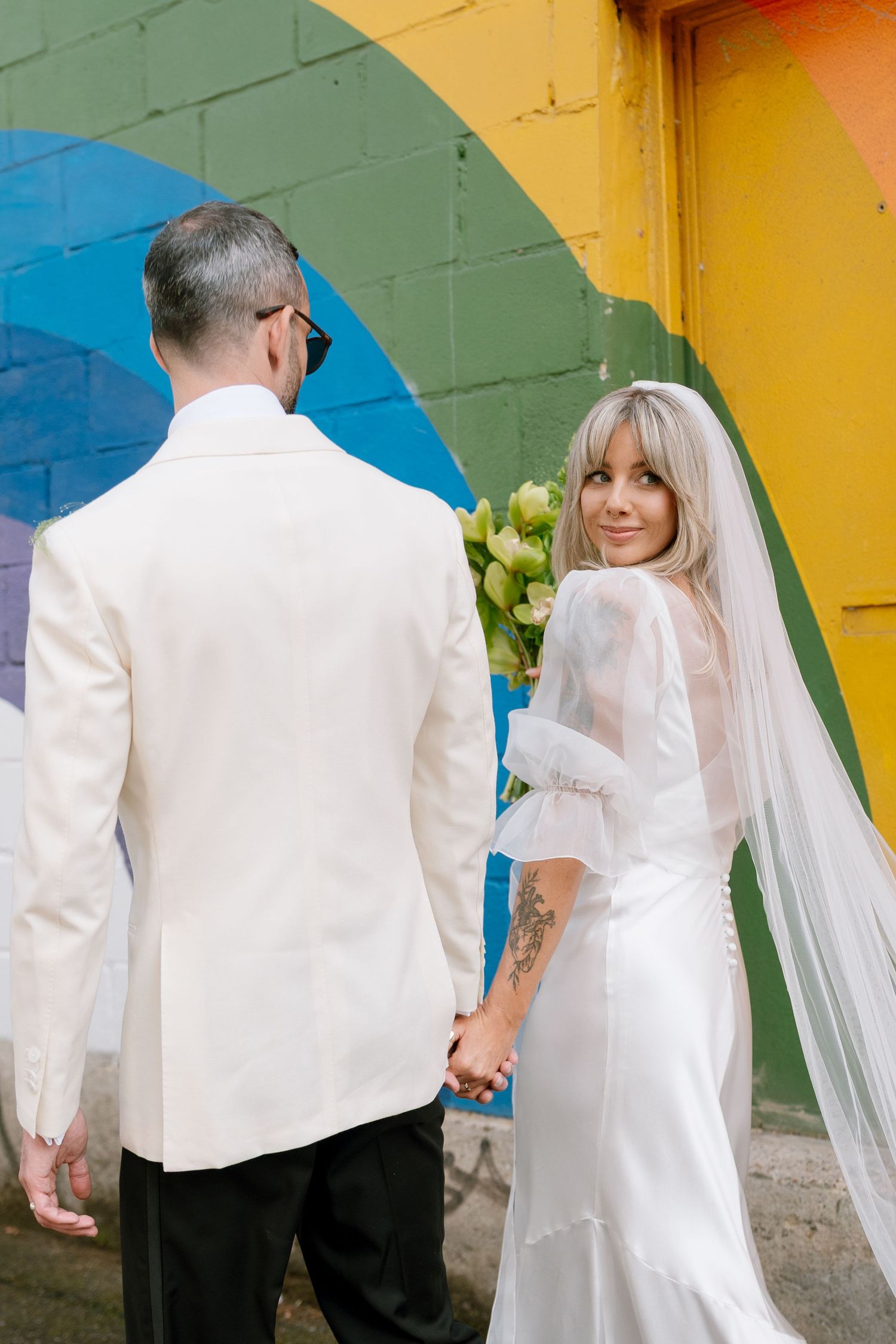 Couple in white wedding attire poses against colorful rainbow wall backdrop.