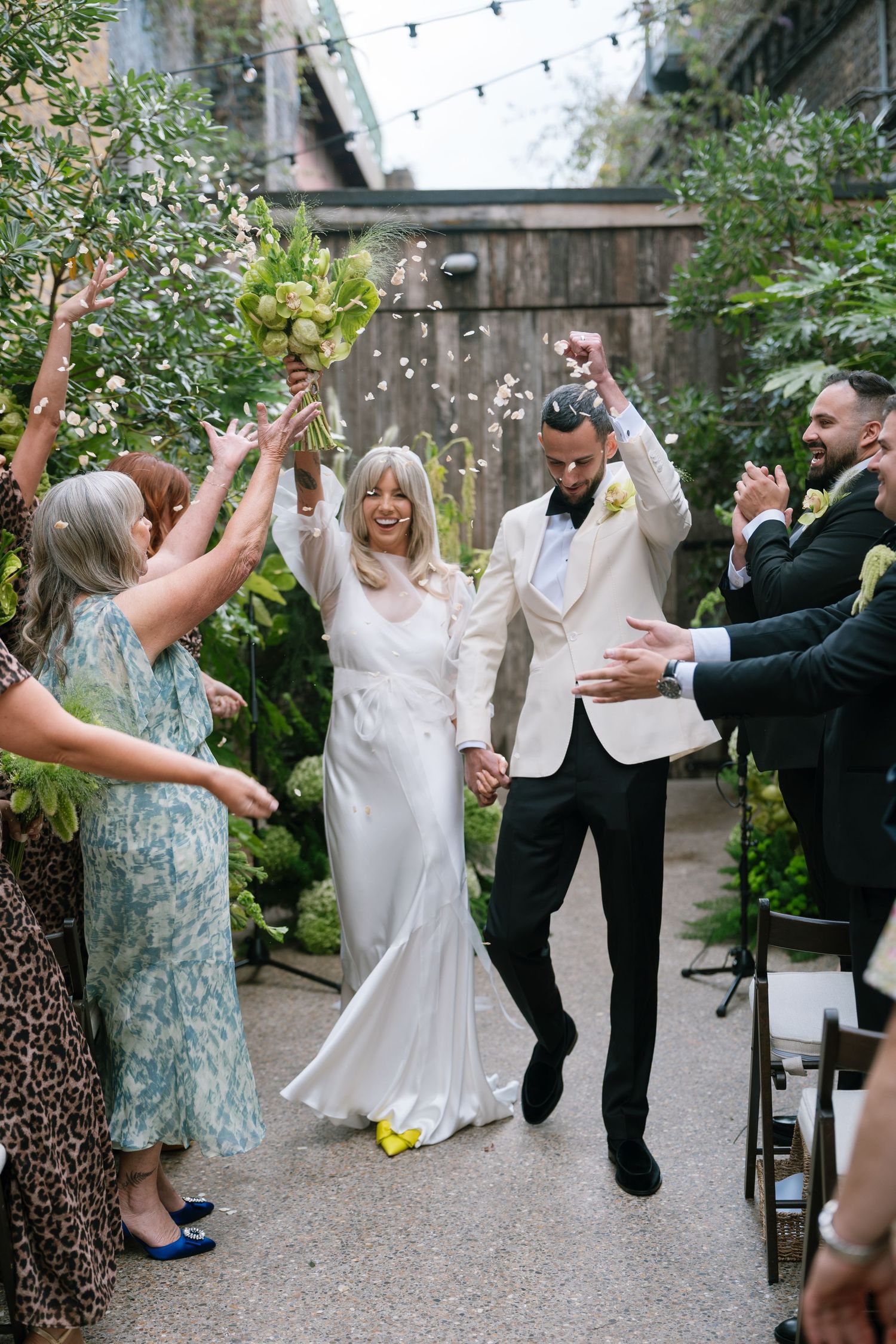 Wedding party dances energetically in an outdoor garden setting with string lights overhead.