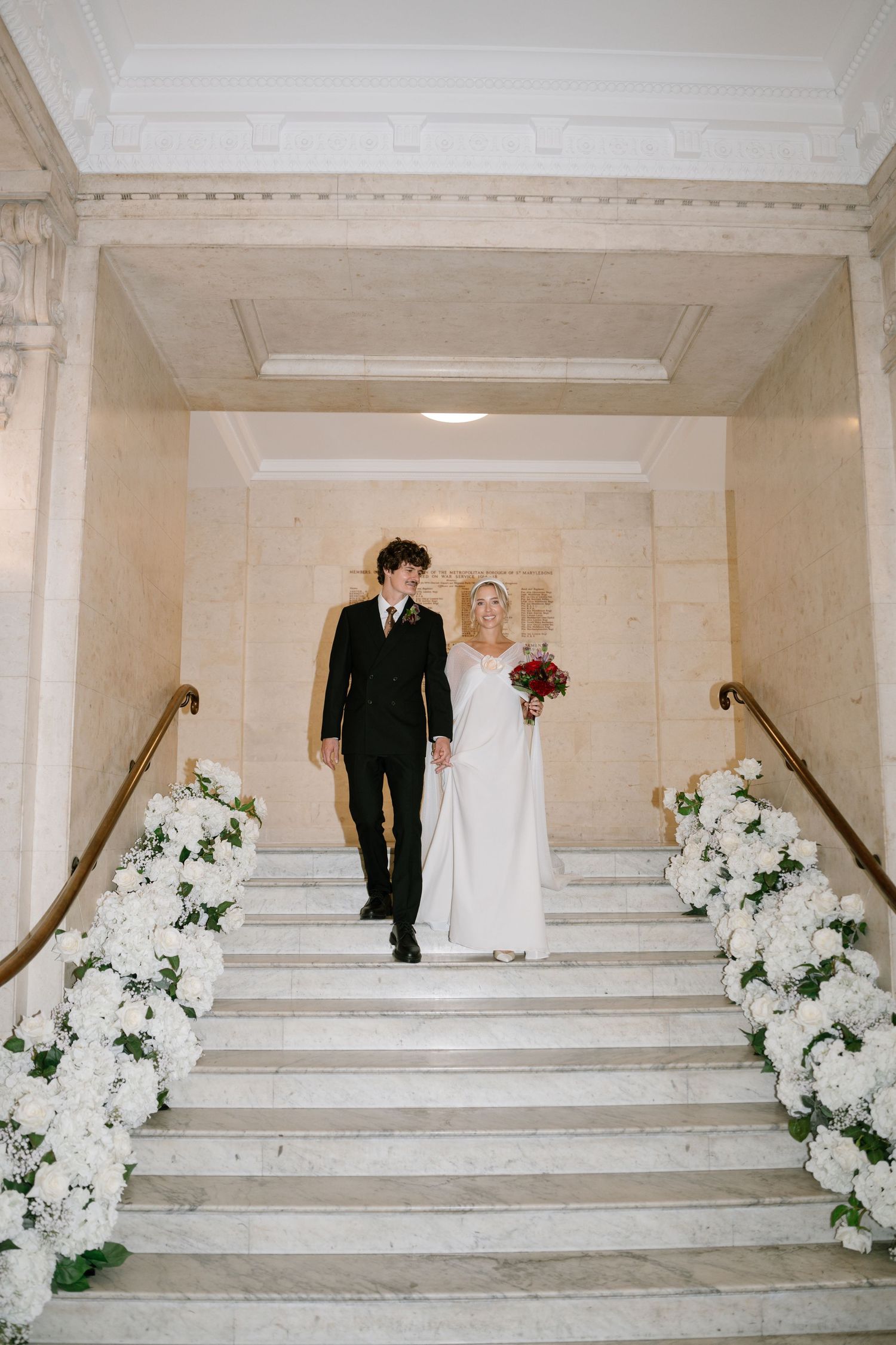 Wedding couple descending white marble stairs decorated with floral arrangements.