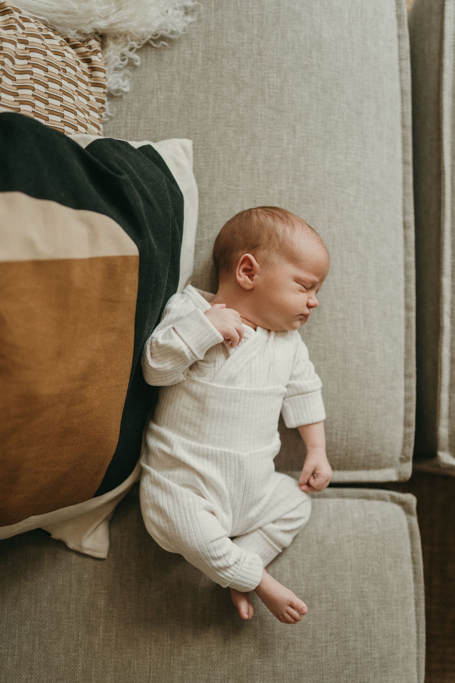 A sleeping infant wearing a white sweater rests peacefully on a gray couch.