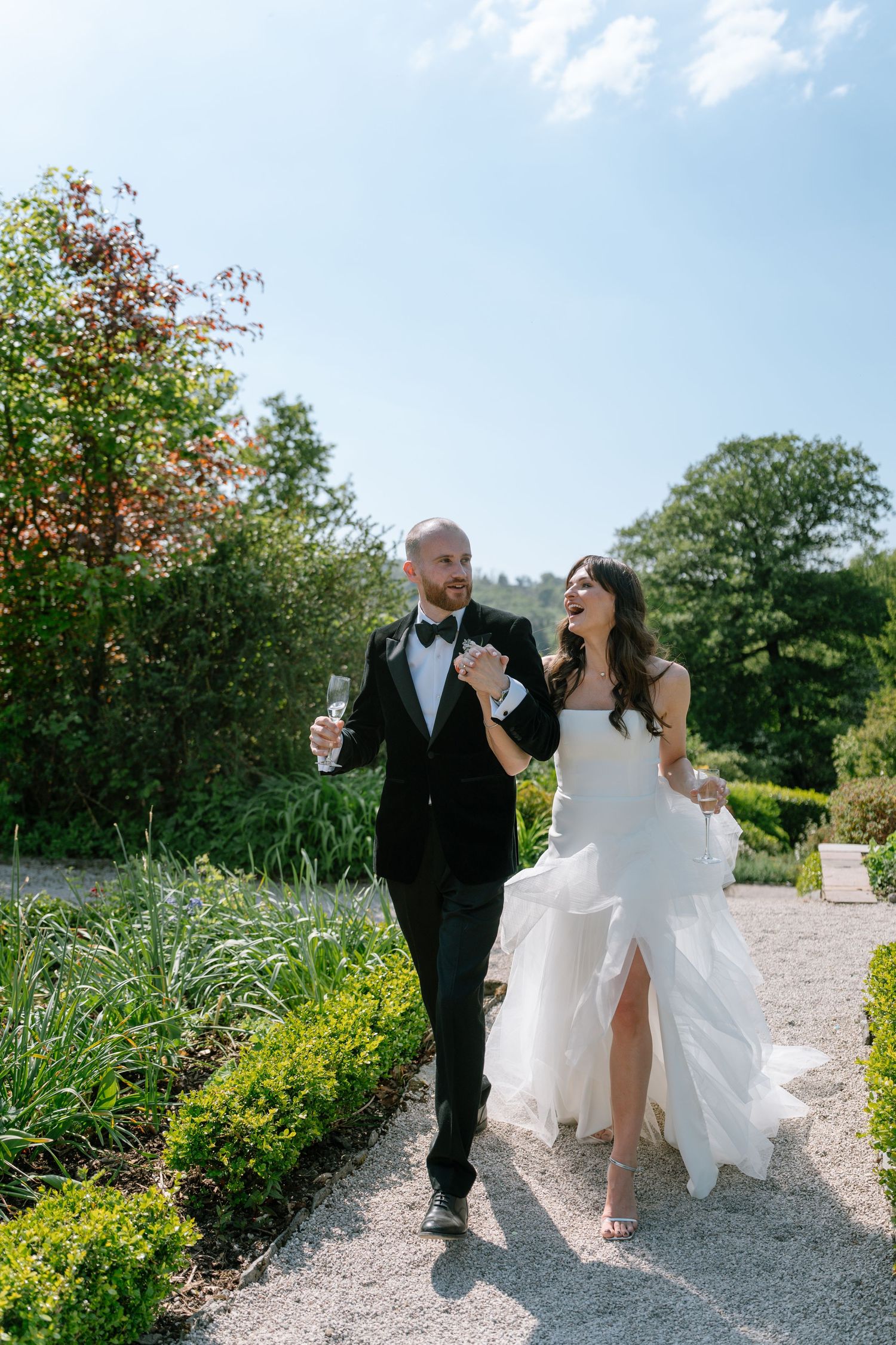 A joyful wedding couple strolls along a gravel garden path on a sunny summer day.