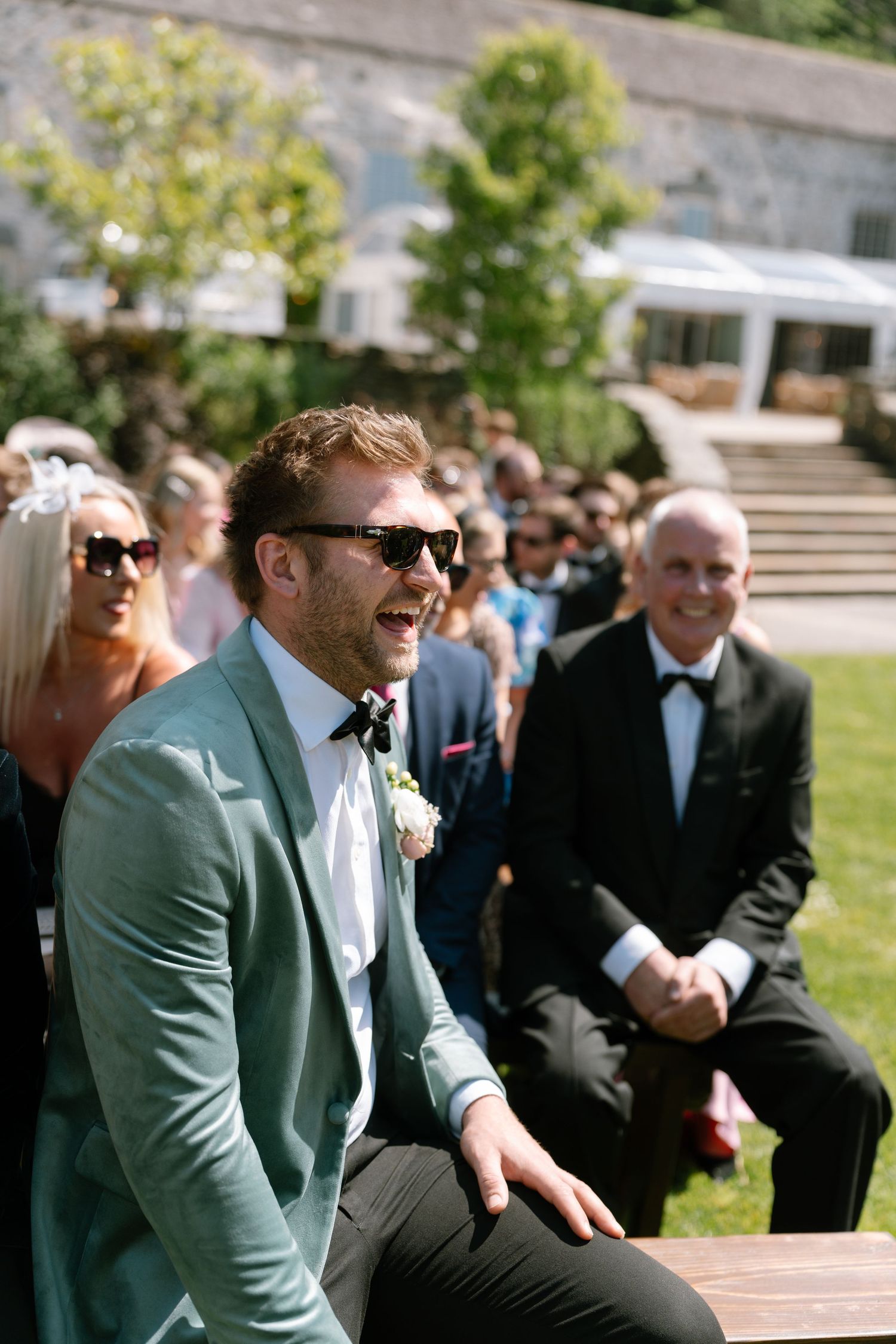 Wedding guests in formal wear and sunglasses enjoy a lighthearted moment in the garden.