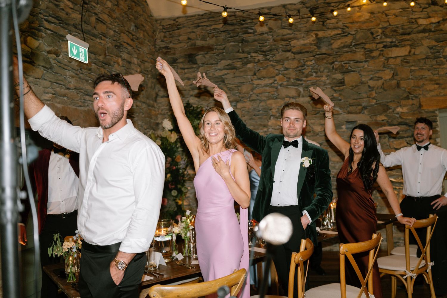 Wedding guests dancing with arms raised under rustic venue lighting.