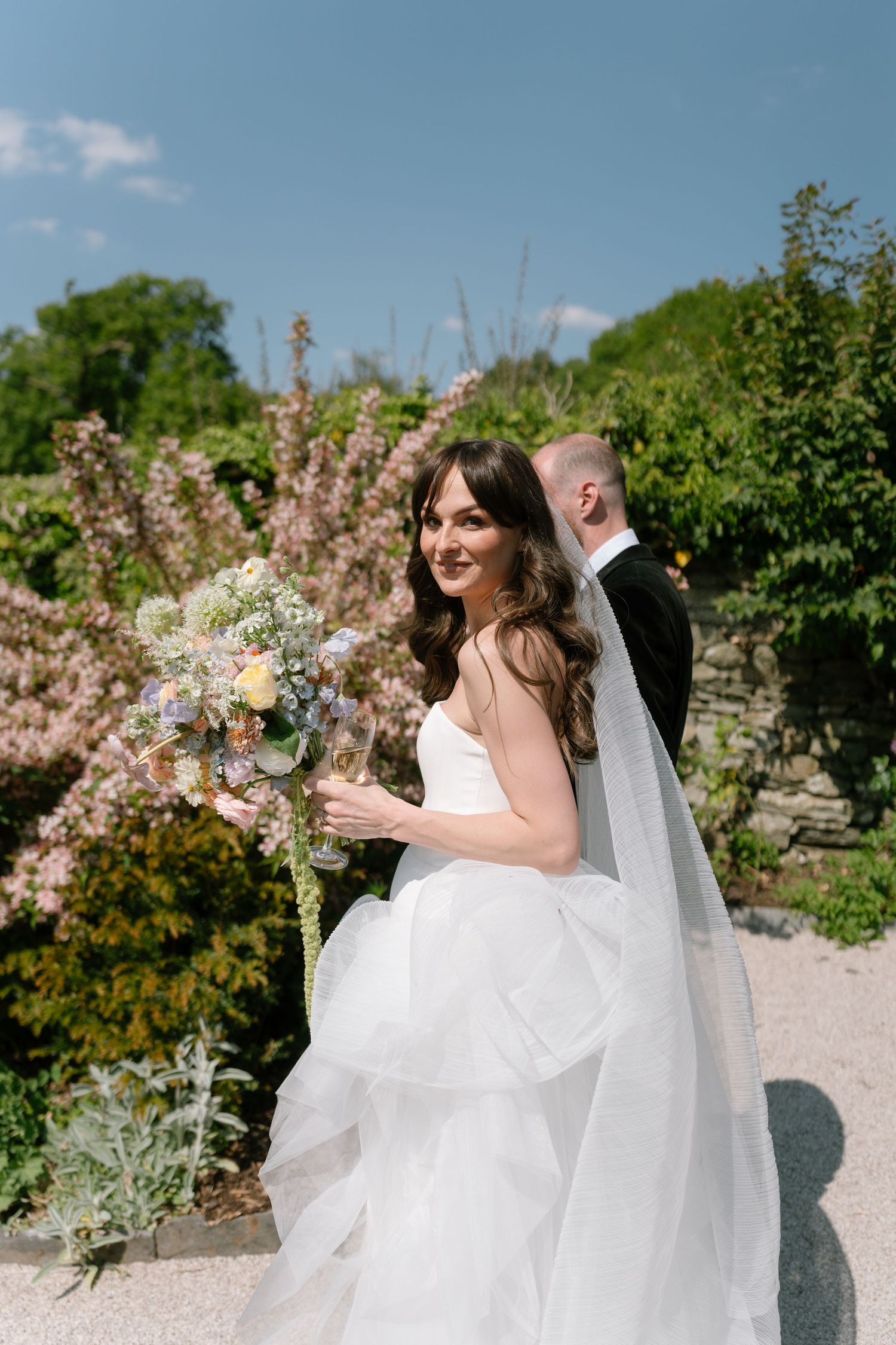 A bride in a flowing white dress walks through a garden setting with flowering bushes.