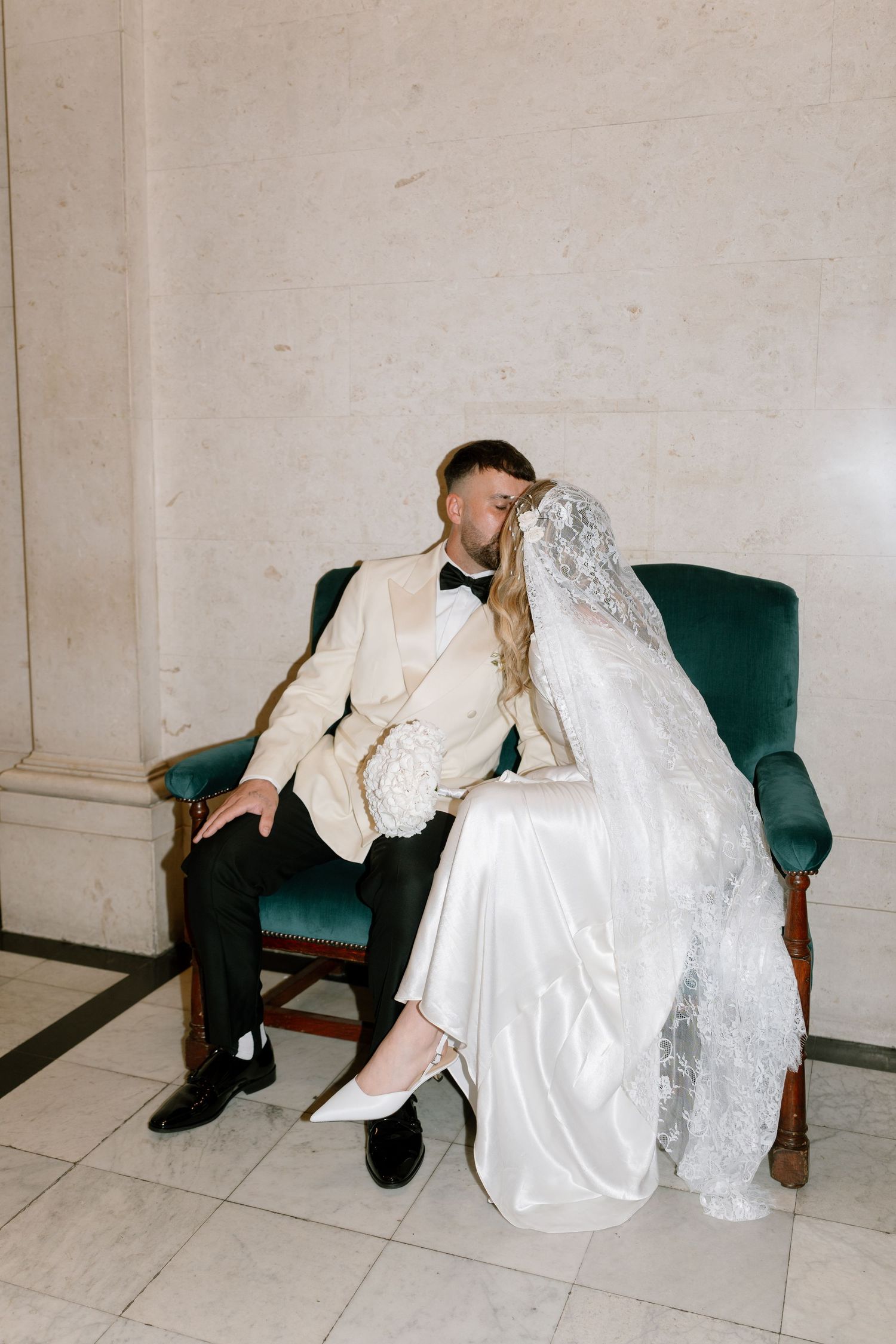 Wedding couple sitting close together on green velvet bench in formal attire.