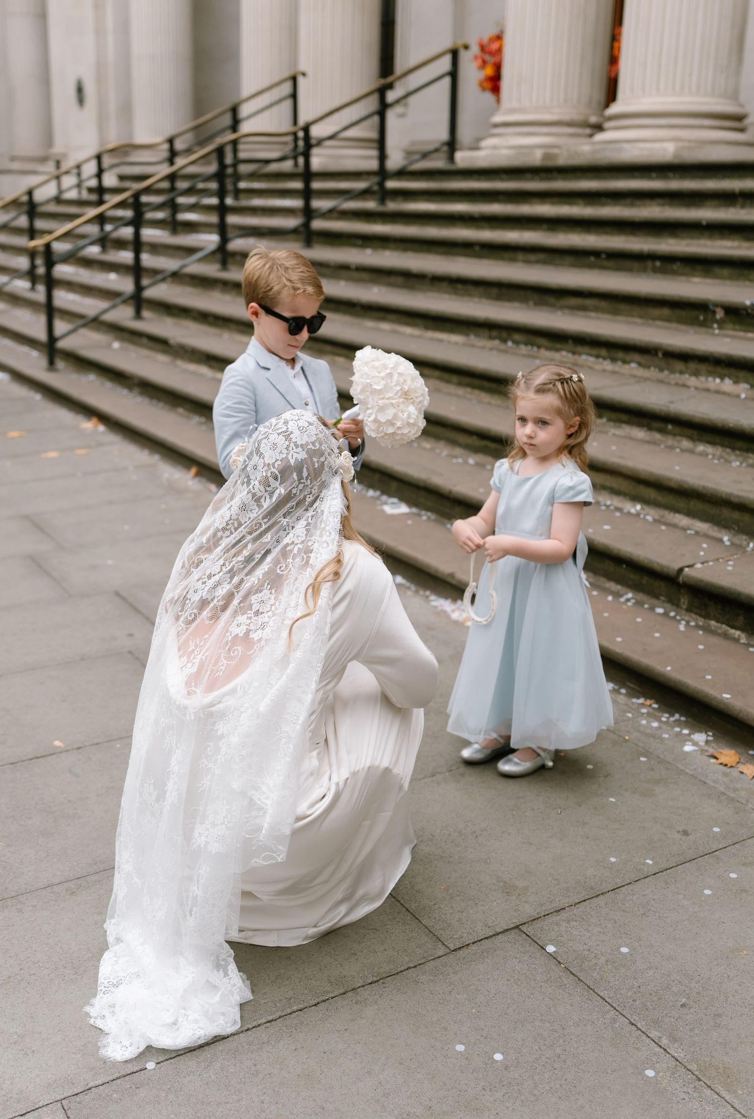 A bride in a lace dress plays with a young flower girl on stone steps outside.