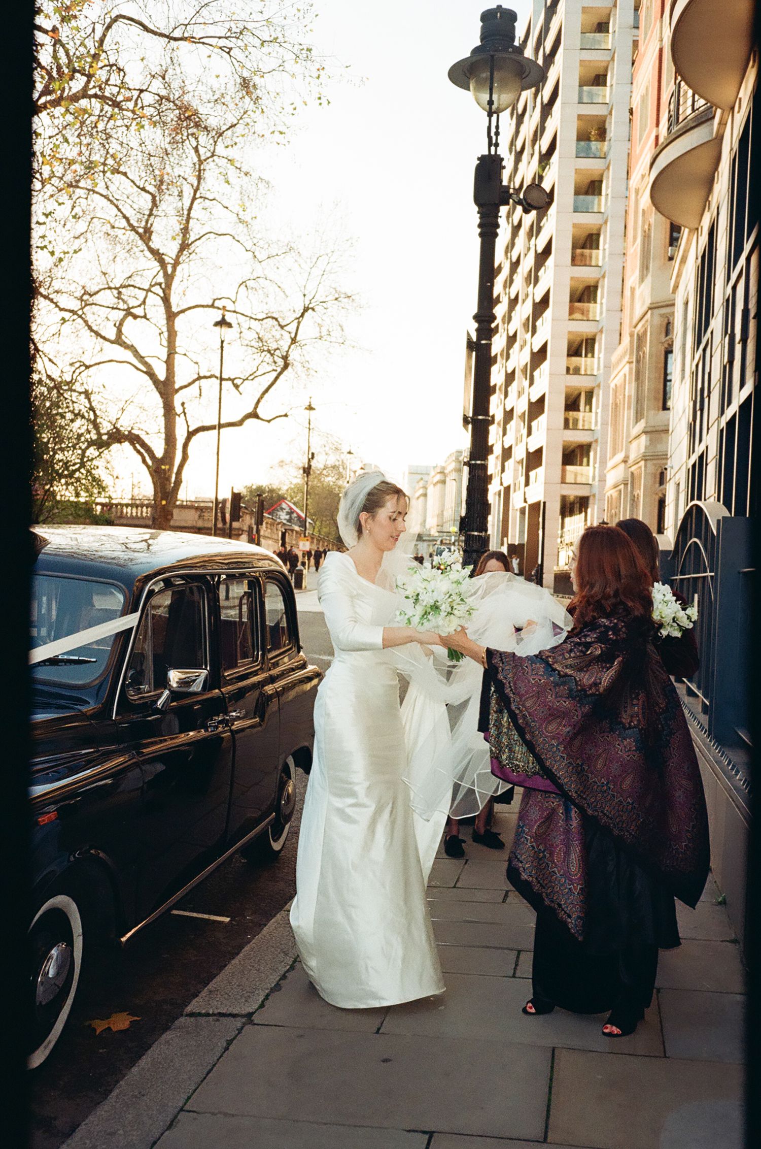 A wedding scene on a city sidewalk with a classic black vintage car and elegant white gown.