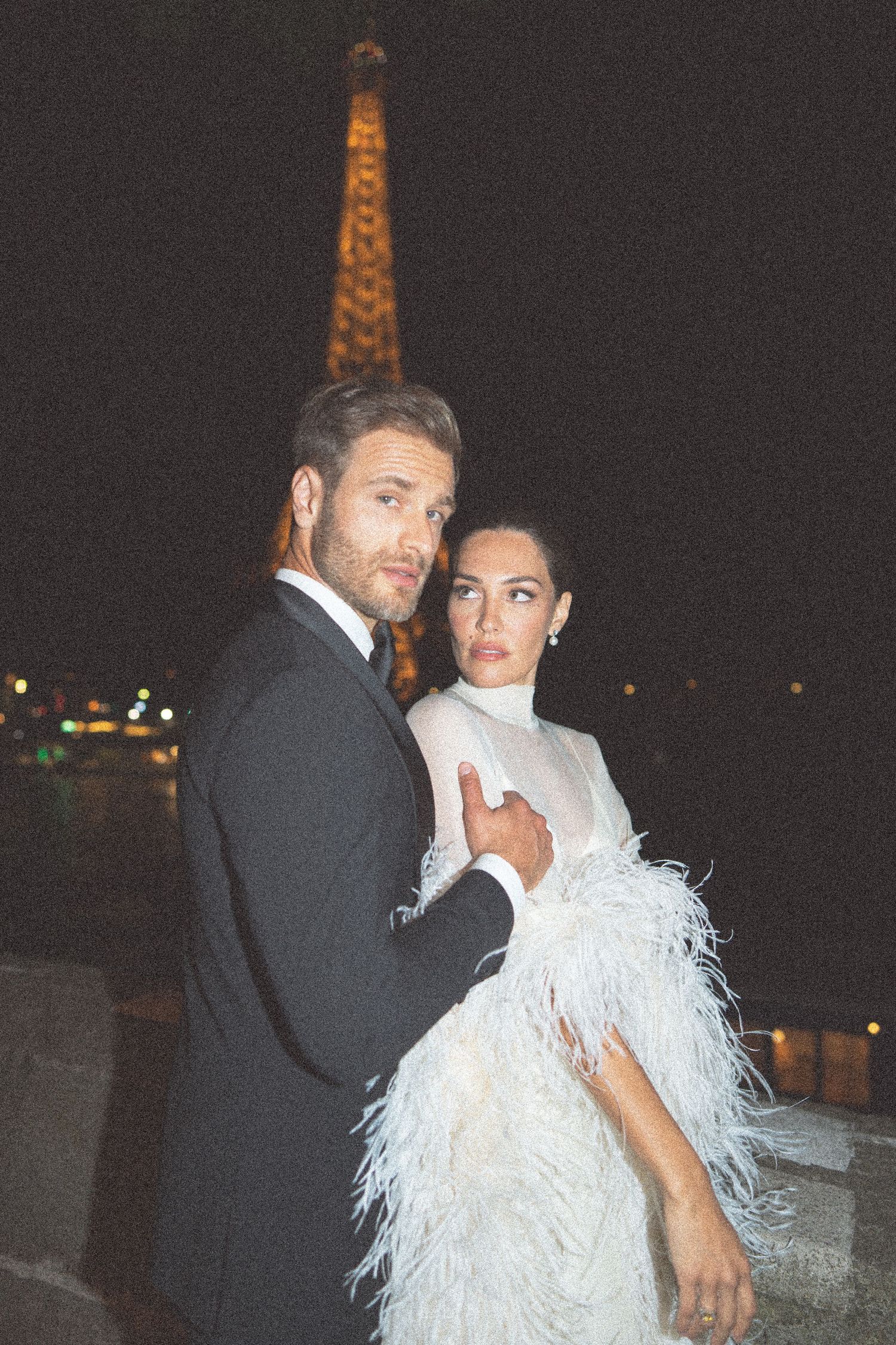 A couple poses in formal attire at night with the illuminated Eiffel Tower visible in the background.