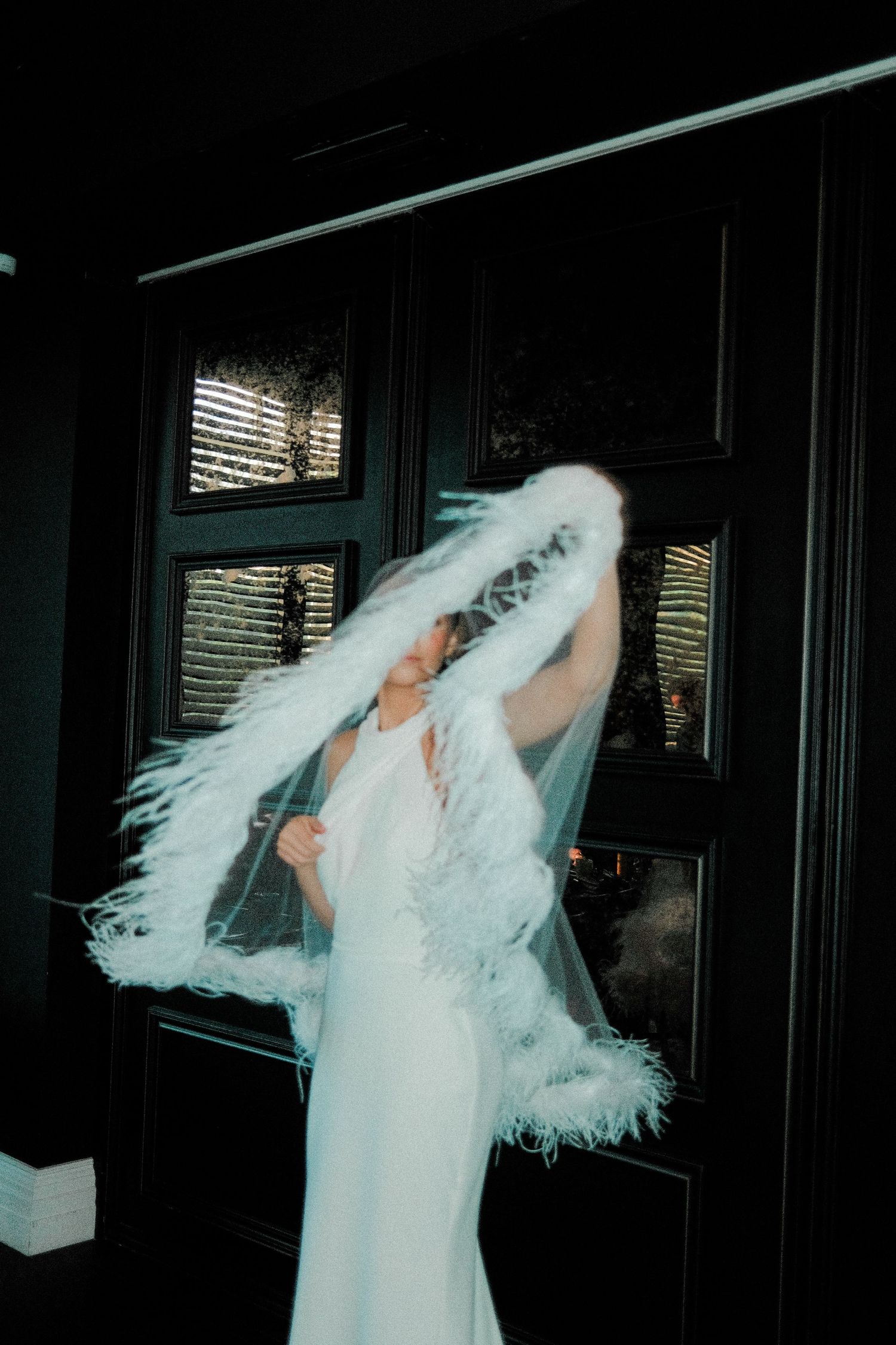 A bride in a flowing white dress and veil twirls gracefully in a dimly lit room with window light streaming in.