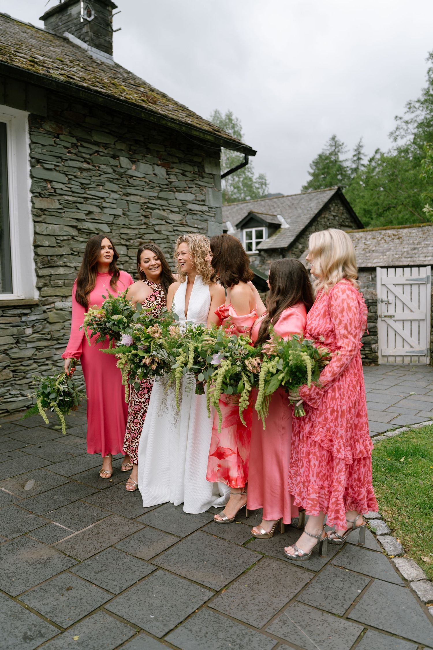 Wedding party in coral pink dresses poses with bride outside stone building.