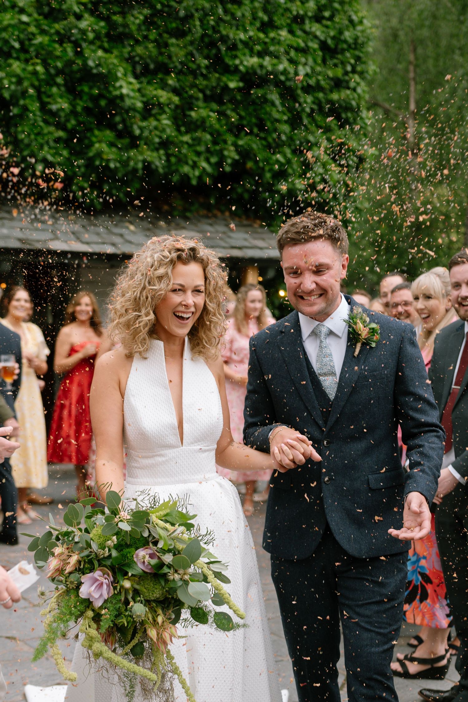 A couple walks through confetti shower outside with greenery in background.