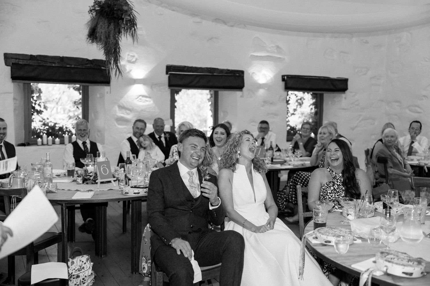 Wedding reception guests seated at tables in rustic venue with exposed beam ceiling.