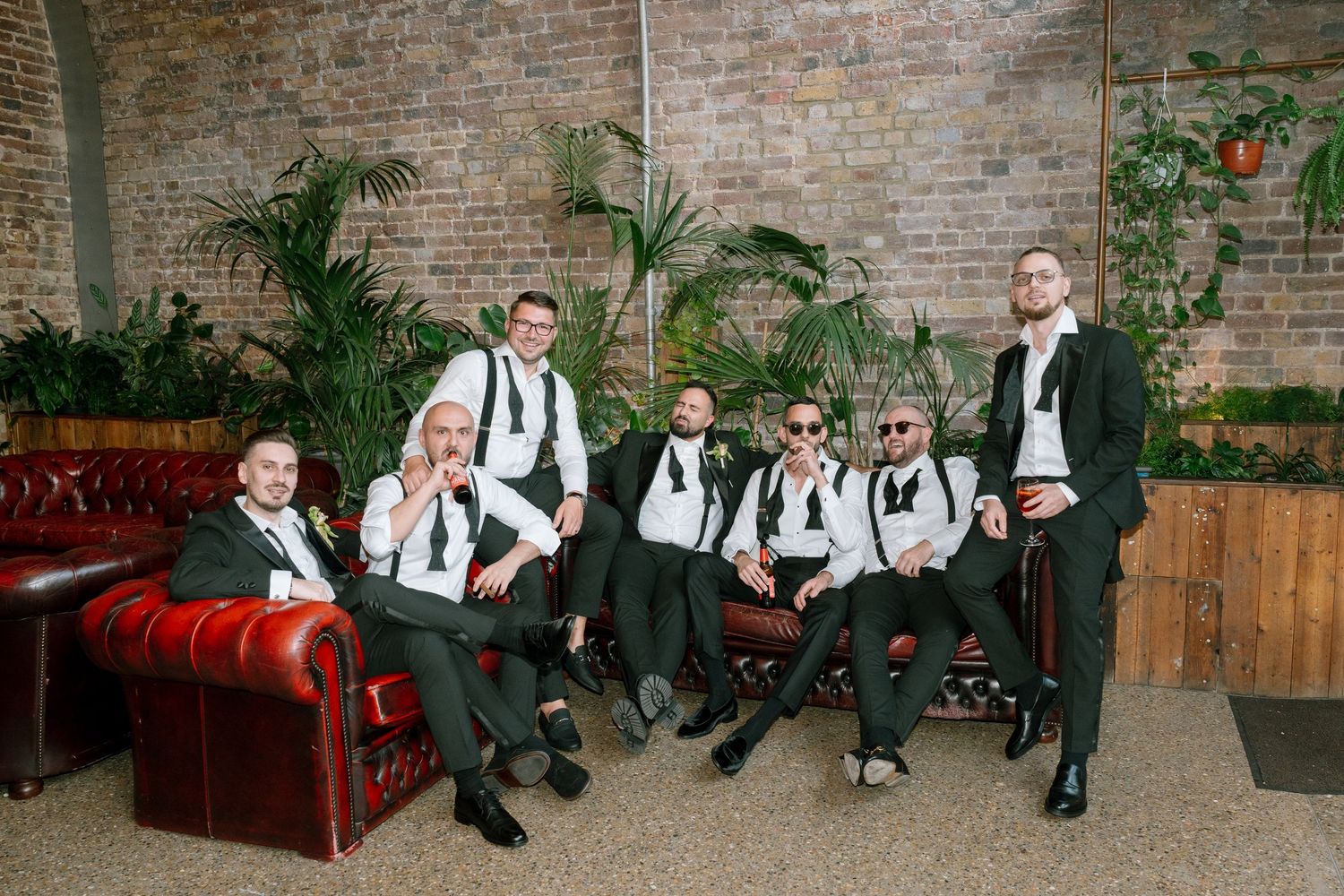 Wedding party in black and white attire poses casually on red leather sofa against brick wall.