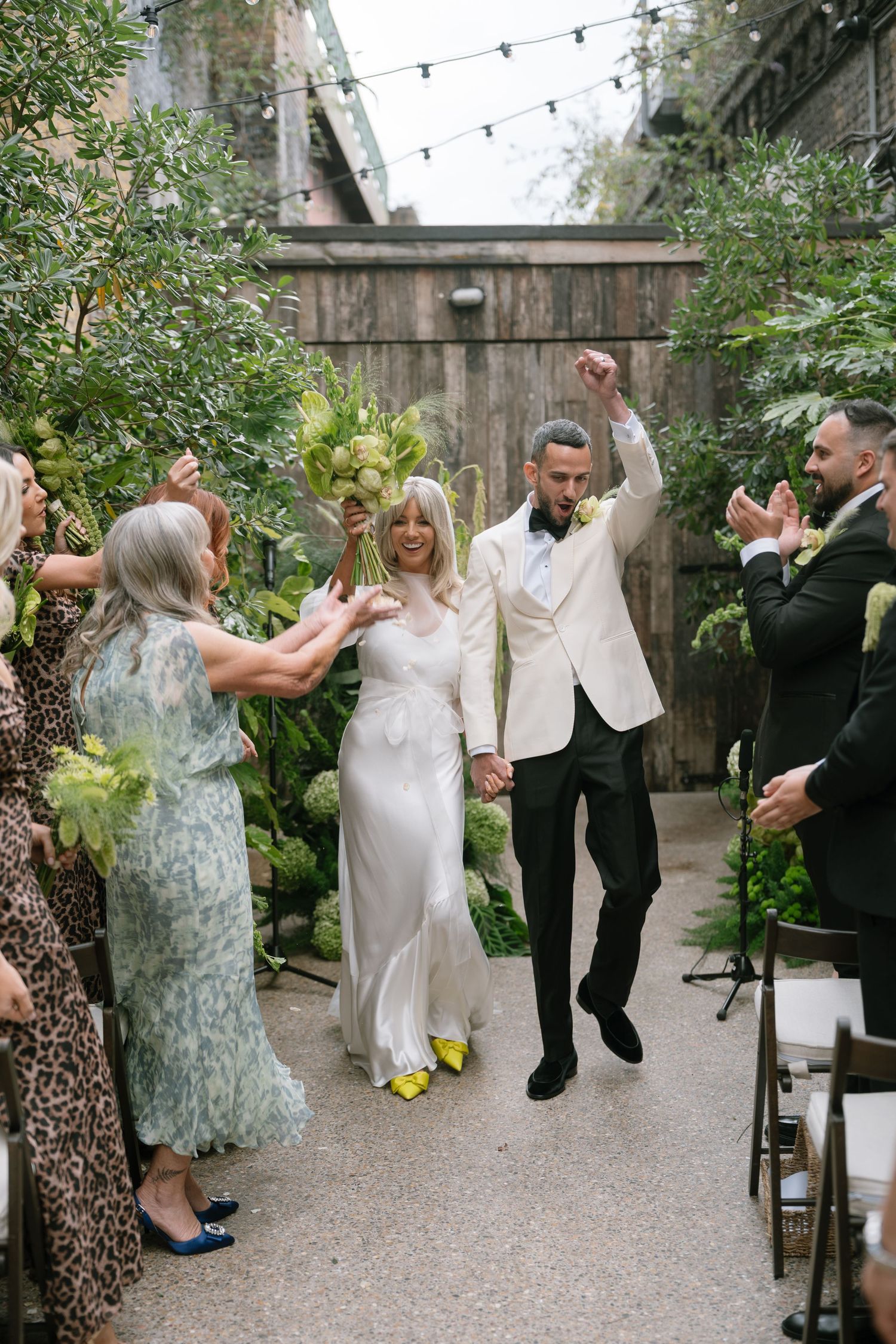 Wedding party celebrates joyfully in an outdoor garden setting with string lights overhead.