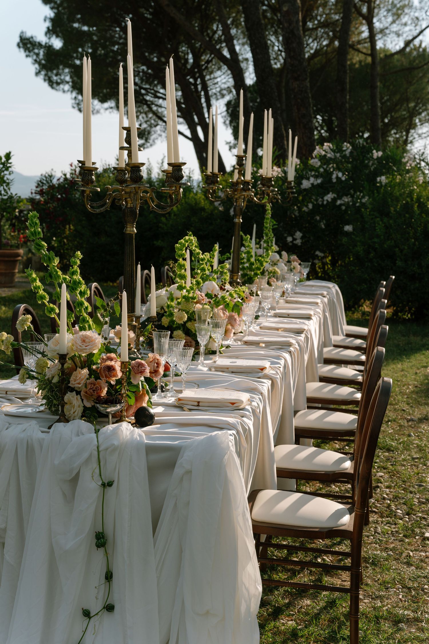 Long outdoor dining table with white linens and floral centerpieces set under tall trees.