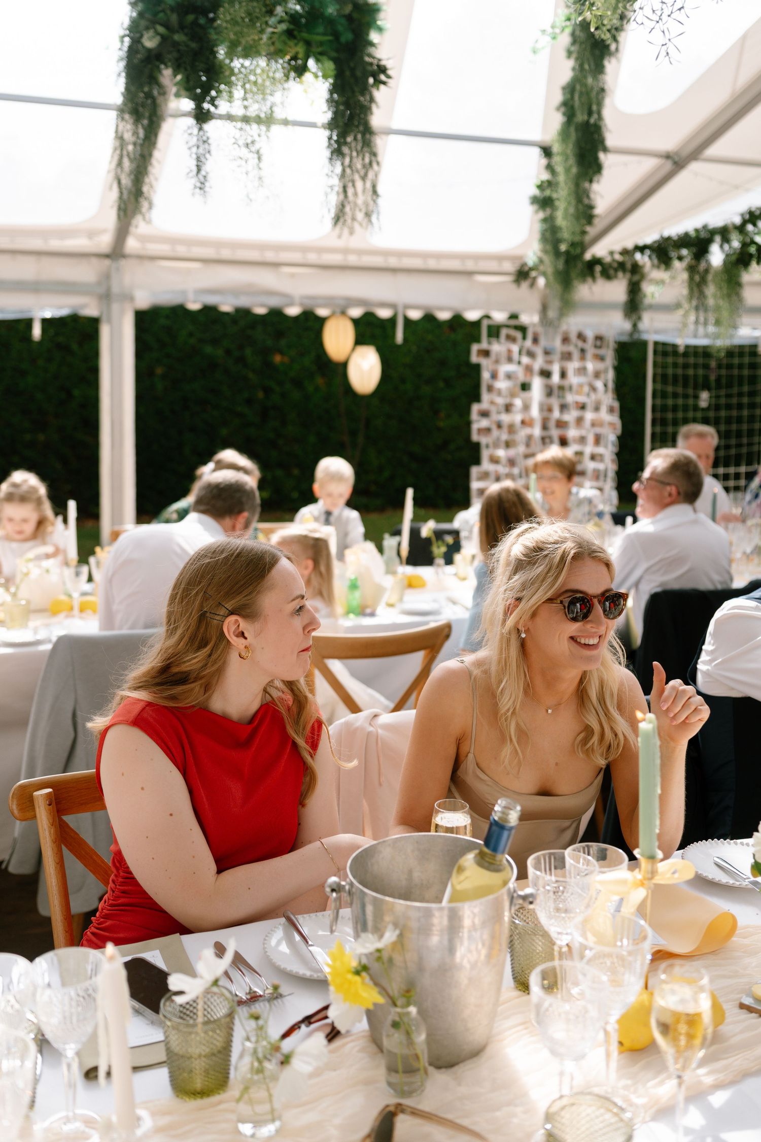Friends laughing together at an outdoor wedding reception under a tent with hanging greenery.