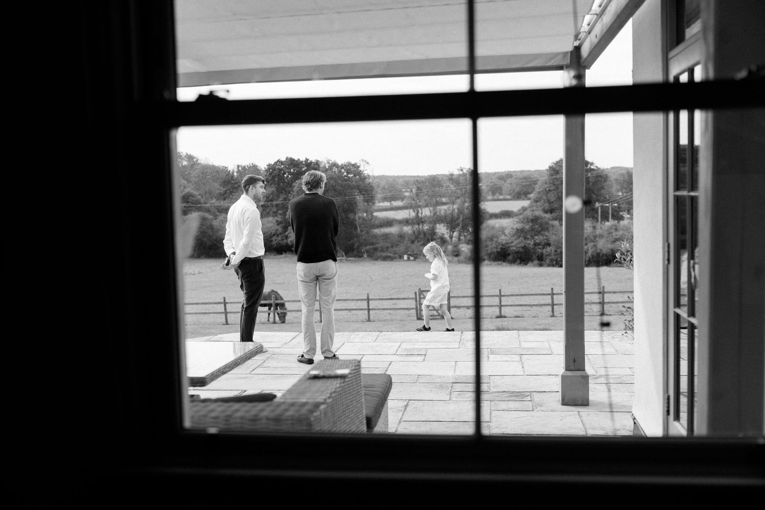 Black and white view through window of people on stone patio with countryside beyond.