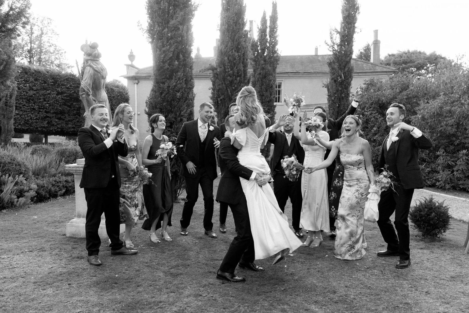 Wedding party walks and poses together on lawn with tall cypress trees creating a natural backdrop.