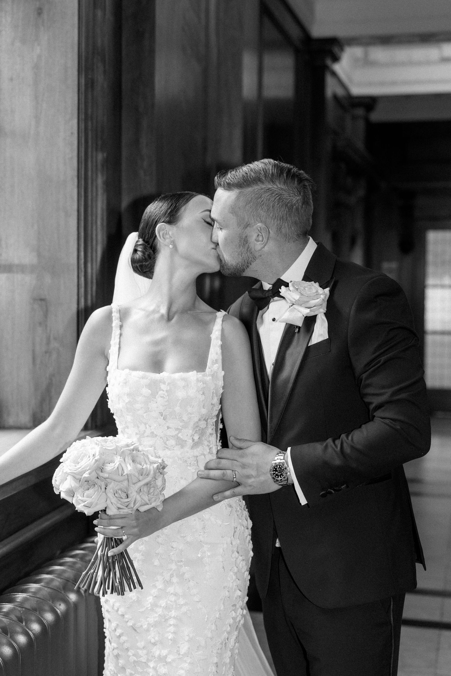Black and white photo of newlyweds kissing near a window in formal attire.