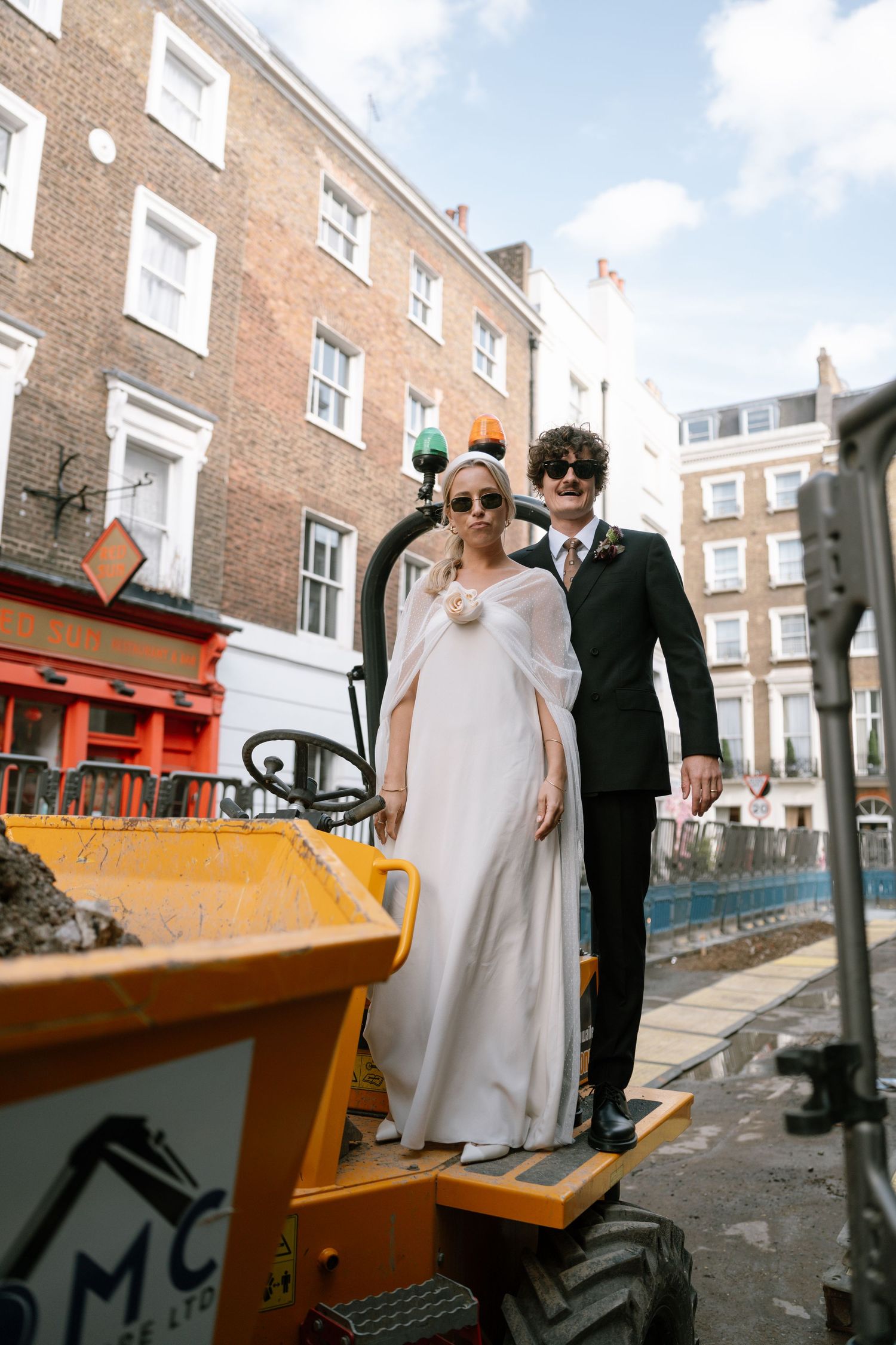 Newlyweds pose together near orange industrial equipment on a sunny urban street.