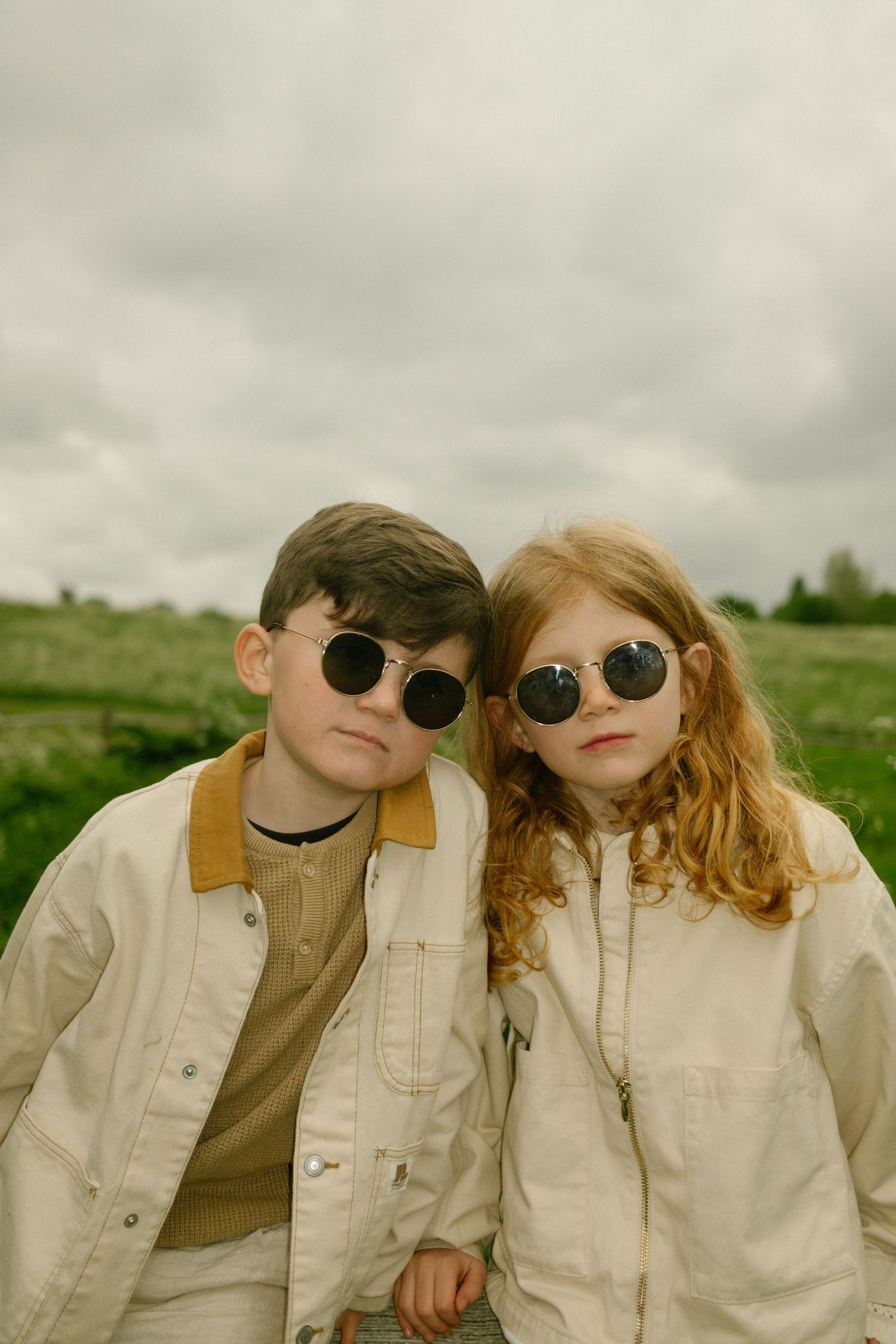Two children in matching white outfits and sunglasses pose playfully in a grassy field.